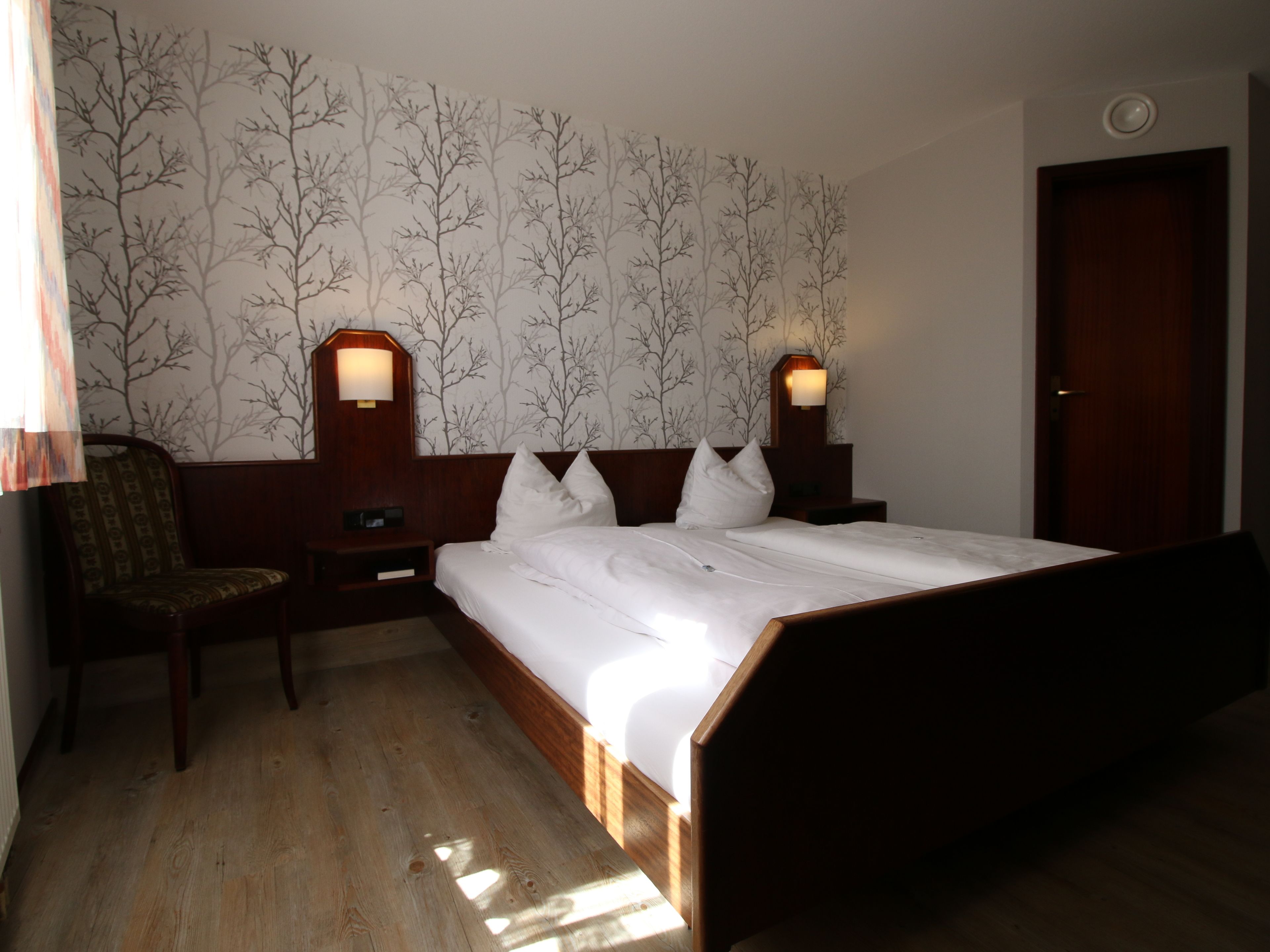 Habitación de hotel con una cama de madera, dos mesitas de noche con lámparas, una silla y un muro con estampado de árboles.