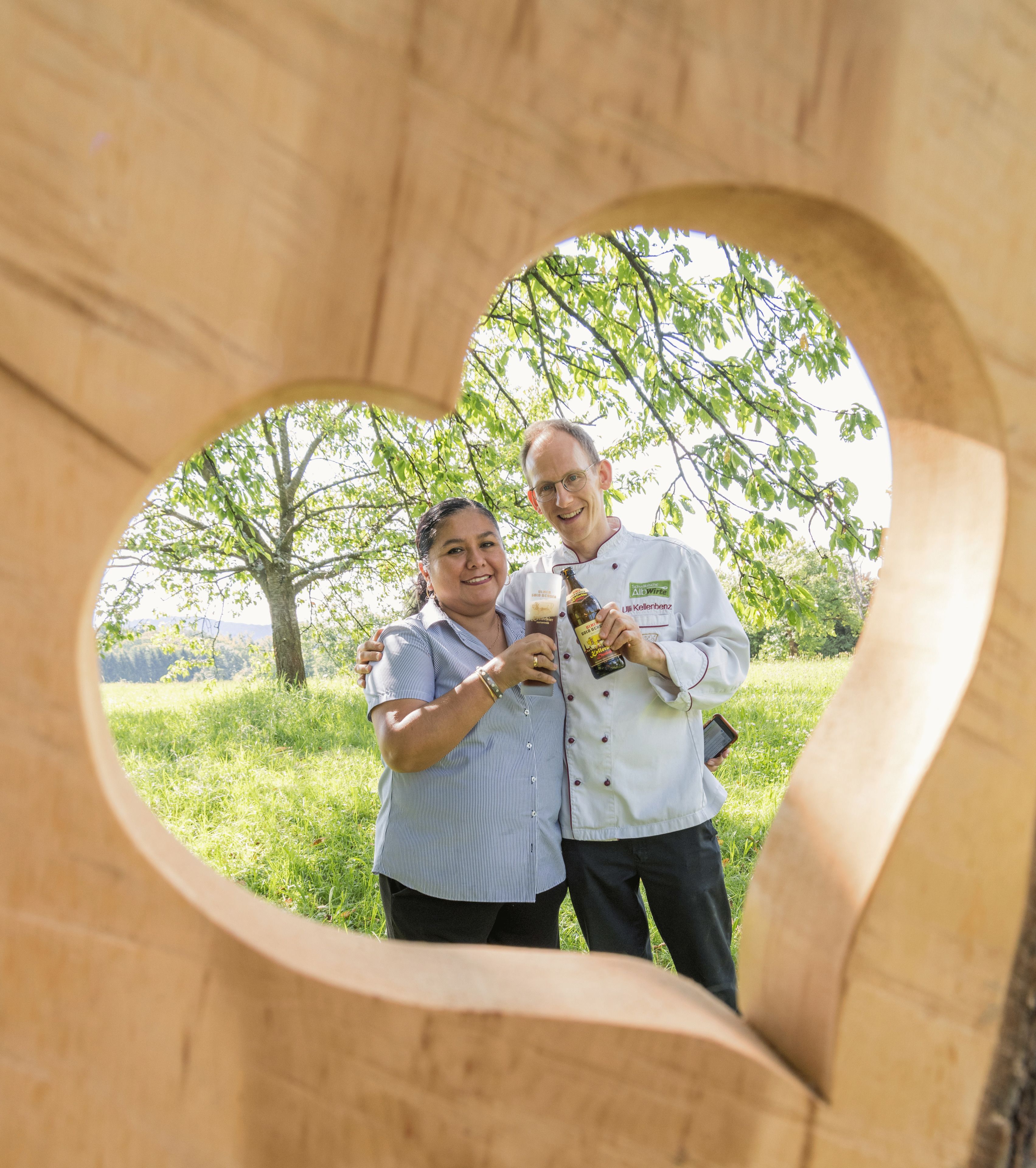 Deux personnes se tiennent debout à l’intérieur d’une structure en bois en forme de cœur, avec un décor naturel en arrière-plan.