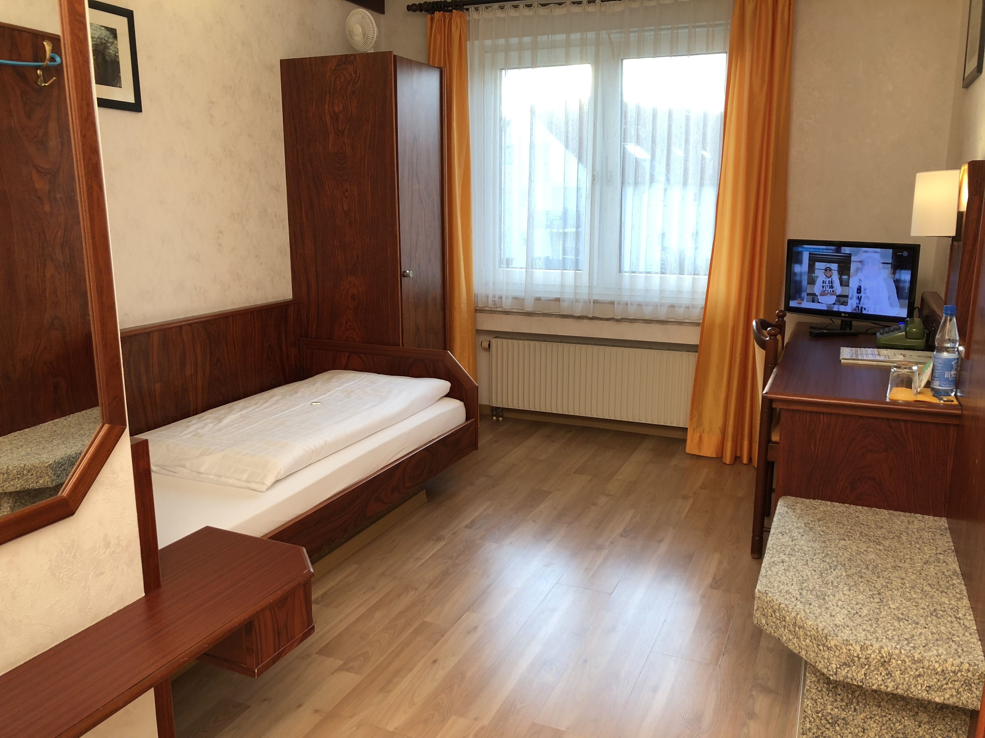 Ein Hotelzimmer mit einem hölzernen Bett, einem Schreibtisch mit Computer, einem Fenster mit Vorhängen und einer Heizung.
