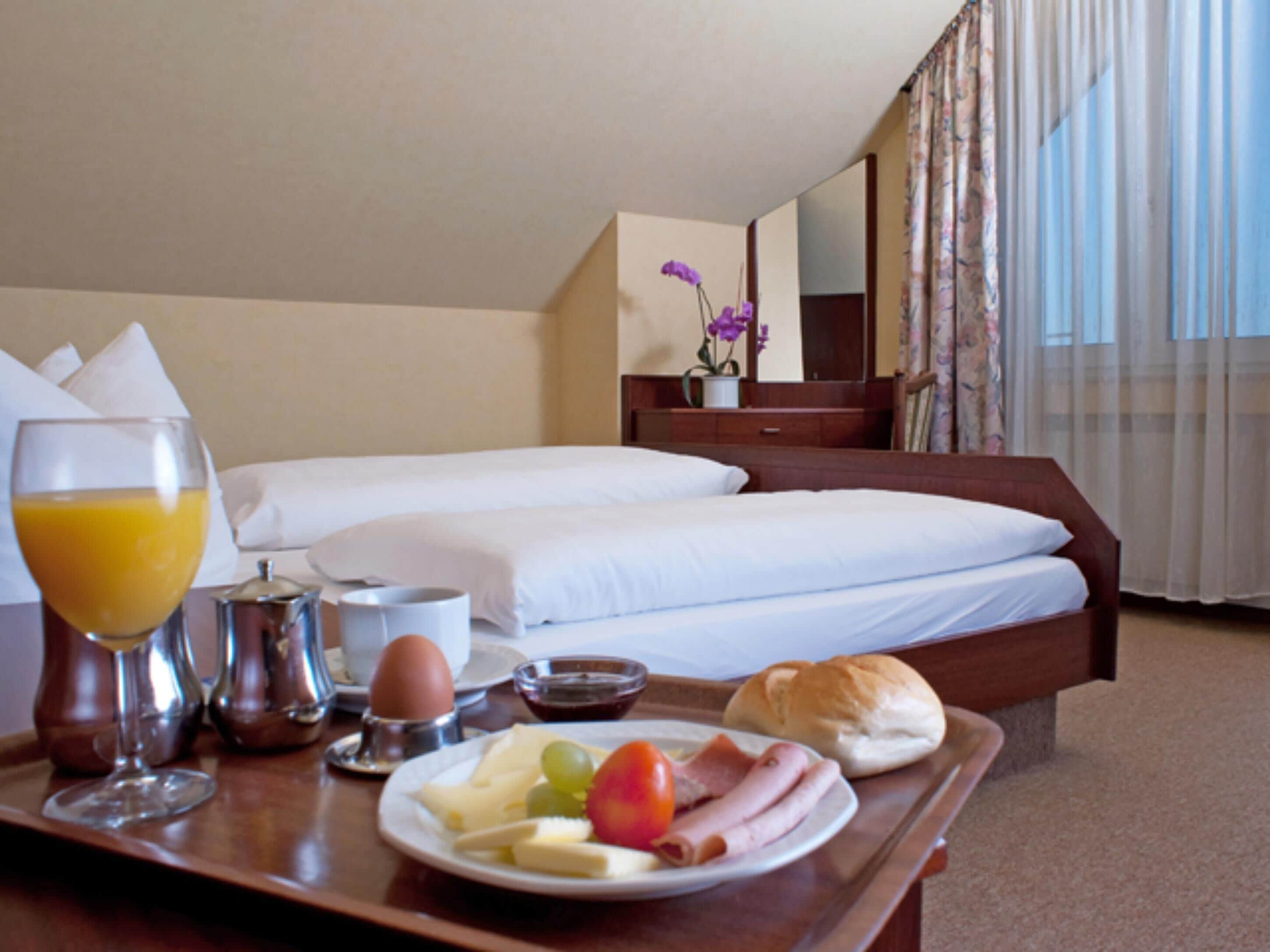Habitación de hotel con cama bien hecha, bandeja de desayuno con huevos, tostadas y fruta, y una ventana con cortinas.