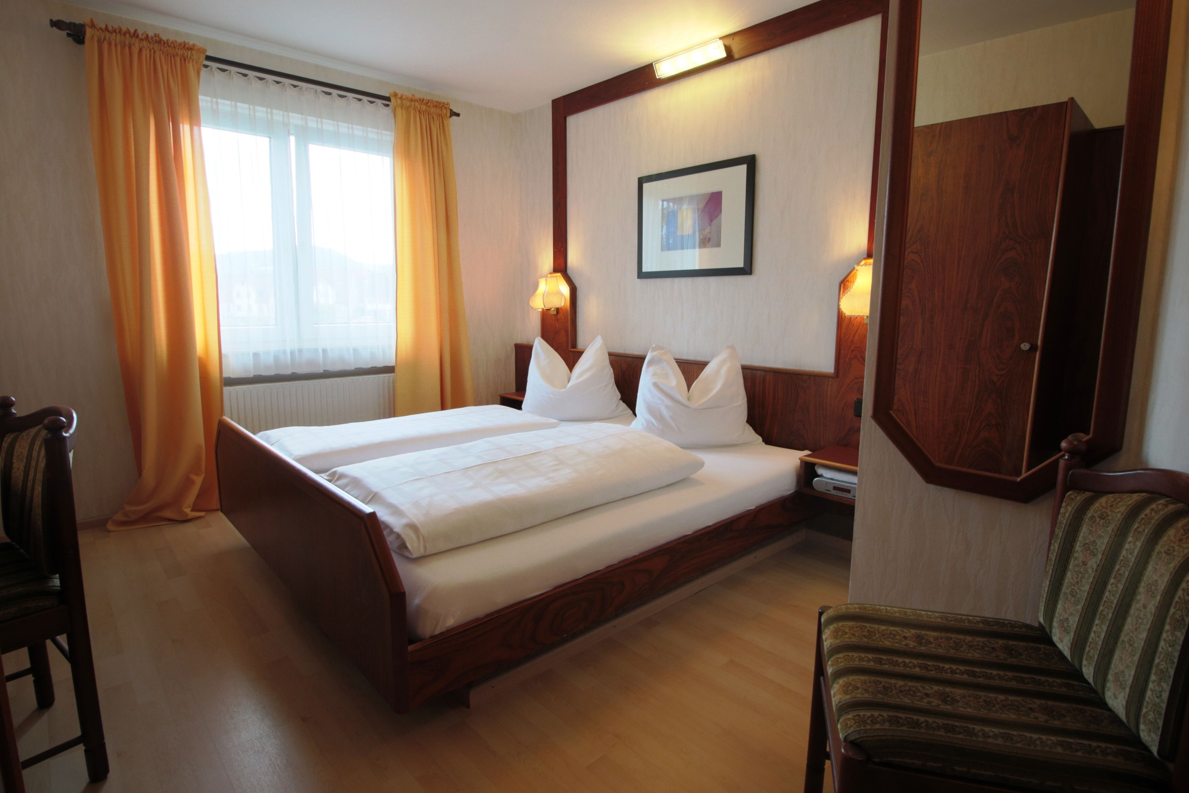 A hotel room with a large bed, two bedside lamps, a wooden wardrobe, a chair, and a window with curtains.