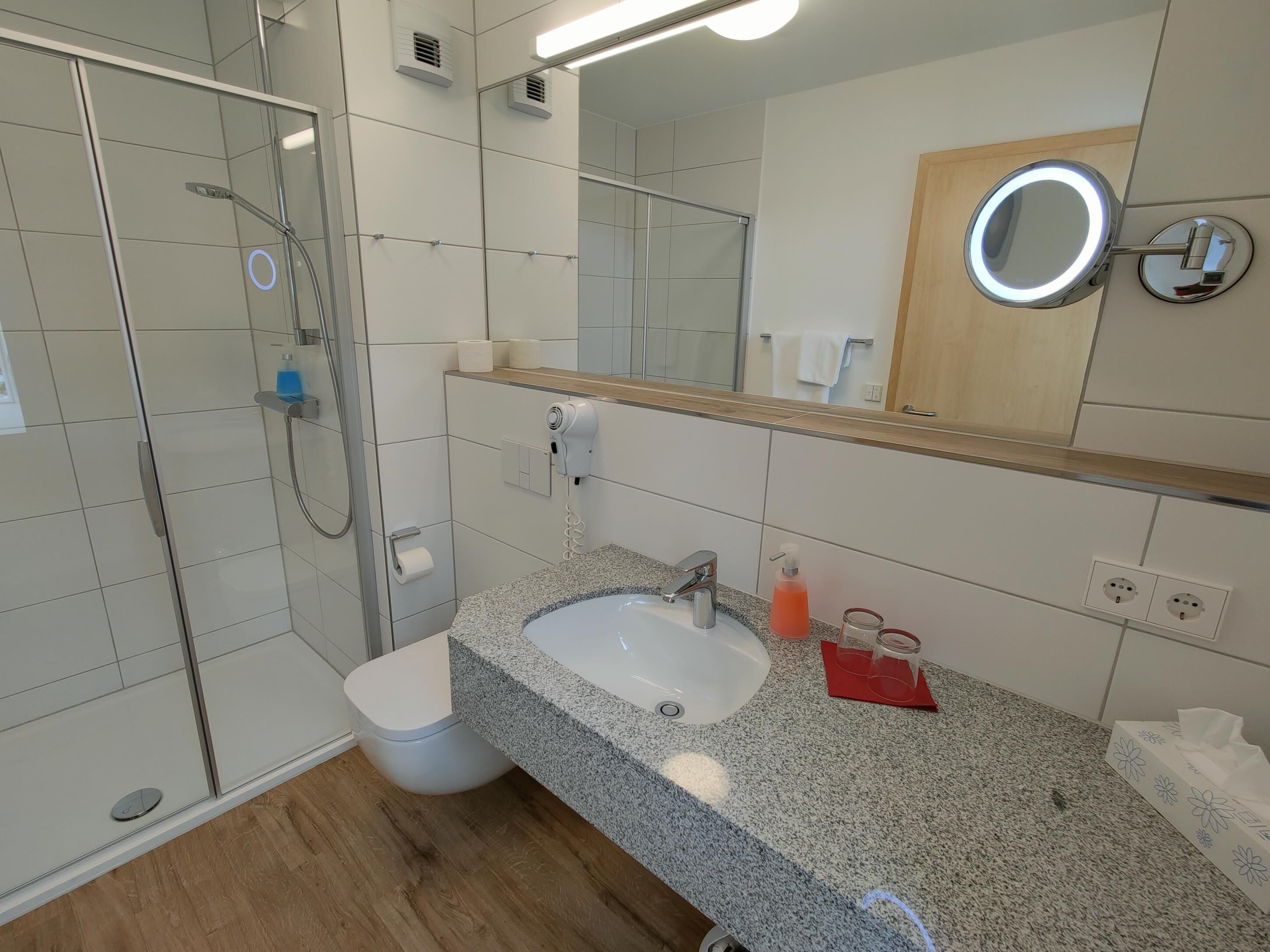 Un baño moderno con cabina de ducha de vidrio, lavamanos con encimera de granito, espejo con luz redonda y tocador de madera.
