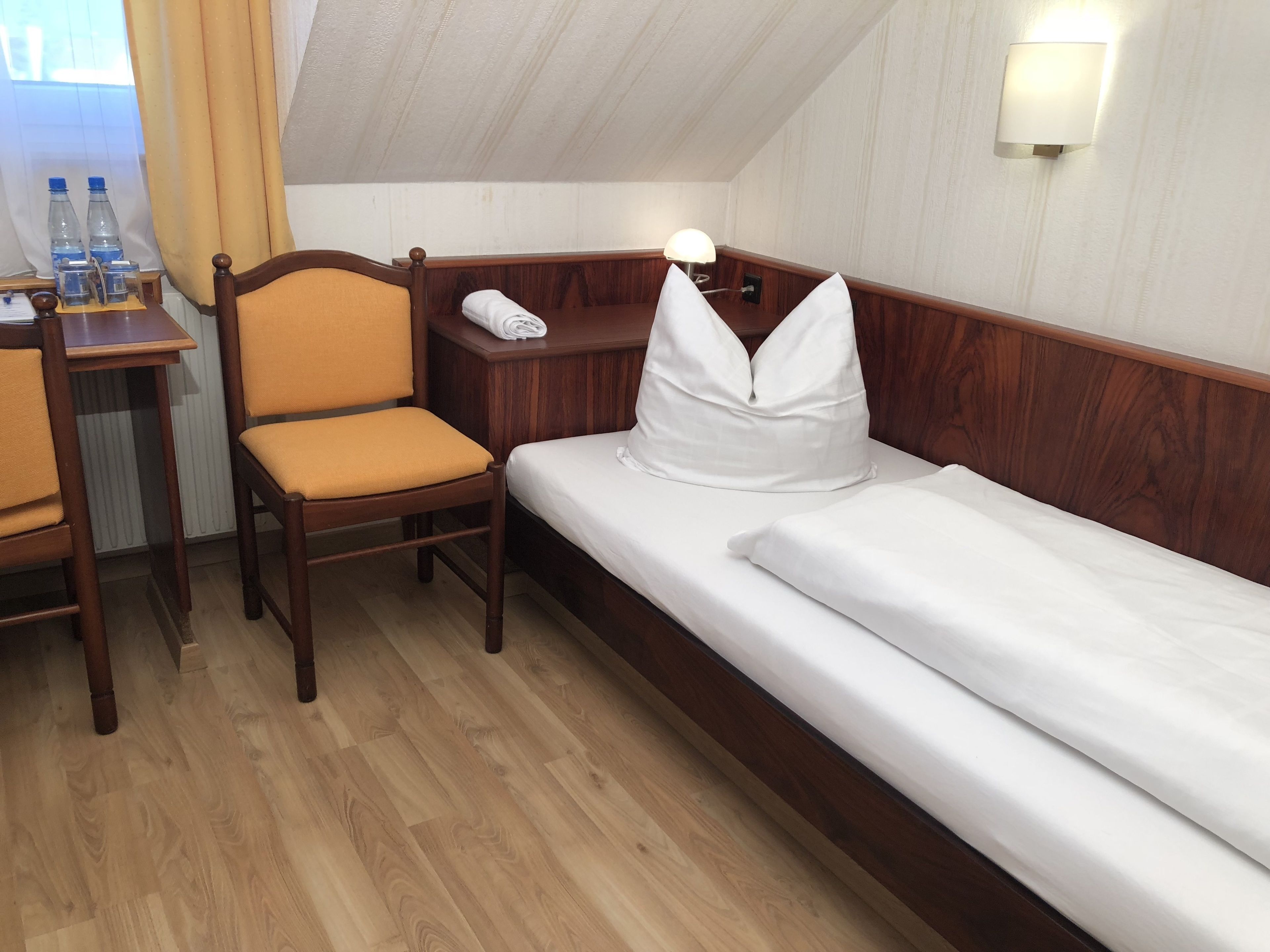 Habitación de hotel con una cama y una silla de madera, una mesita con lámpara y una ventana con cortinas.