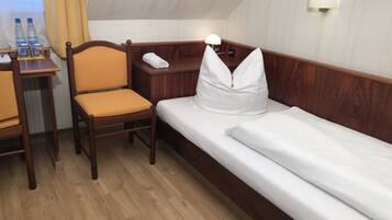 Quarto de hotel com cama e cadeira de madeira, uma mesinha com abajur e uma janela com cortinas.