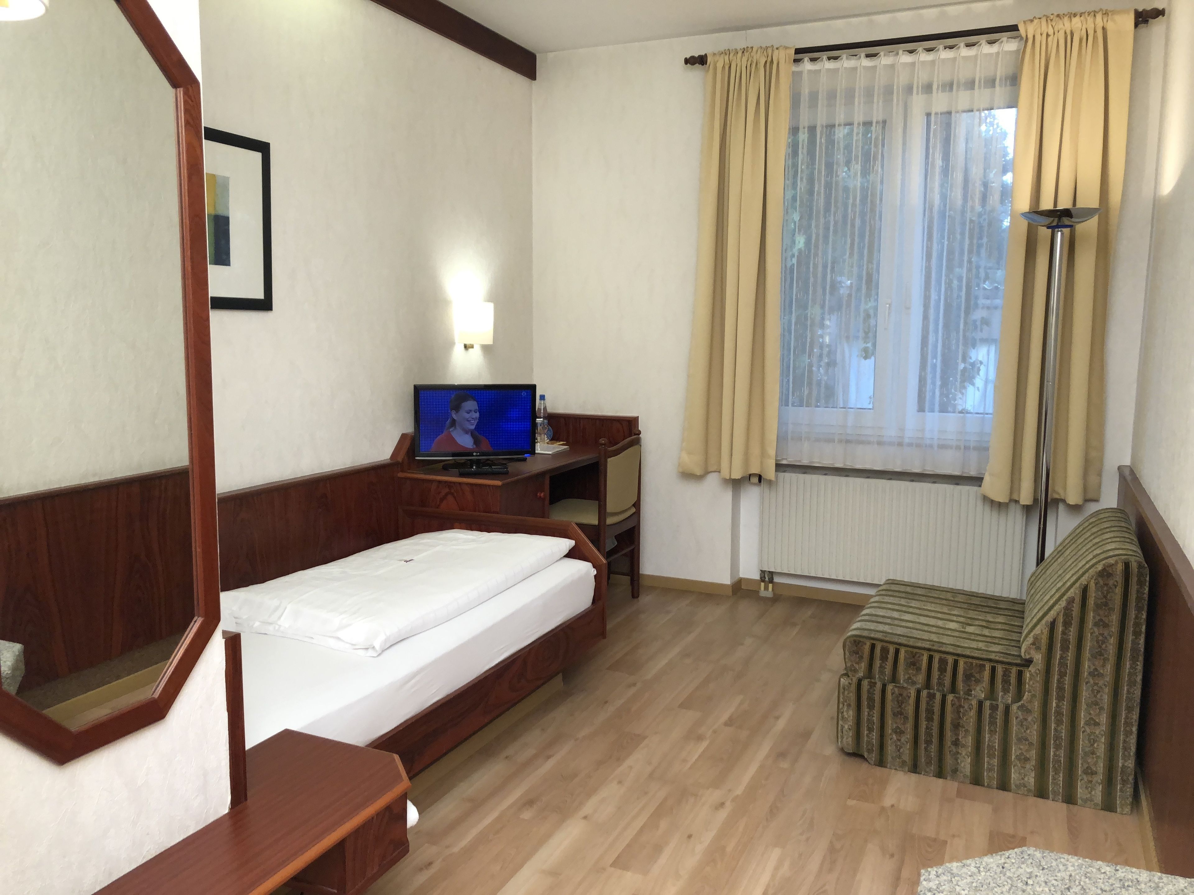 Habitación de hotel con cama, escritorio con computadora, silla, ventana con cortinas y radiador.