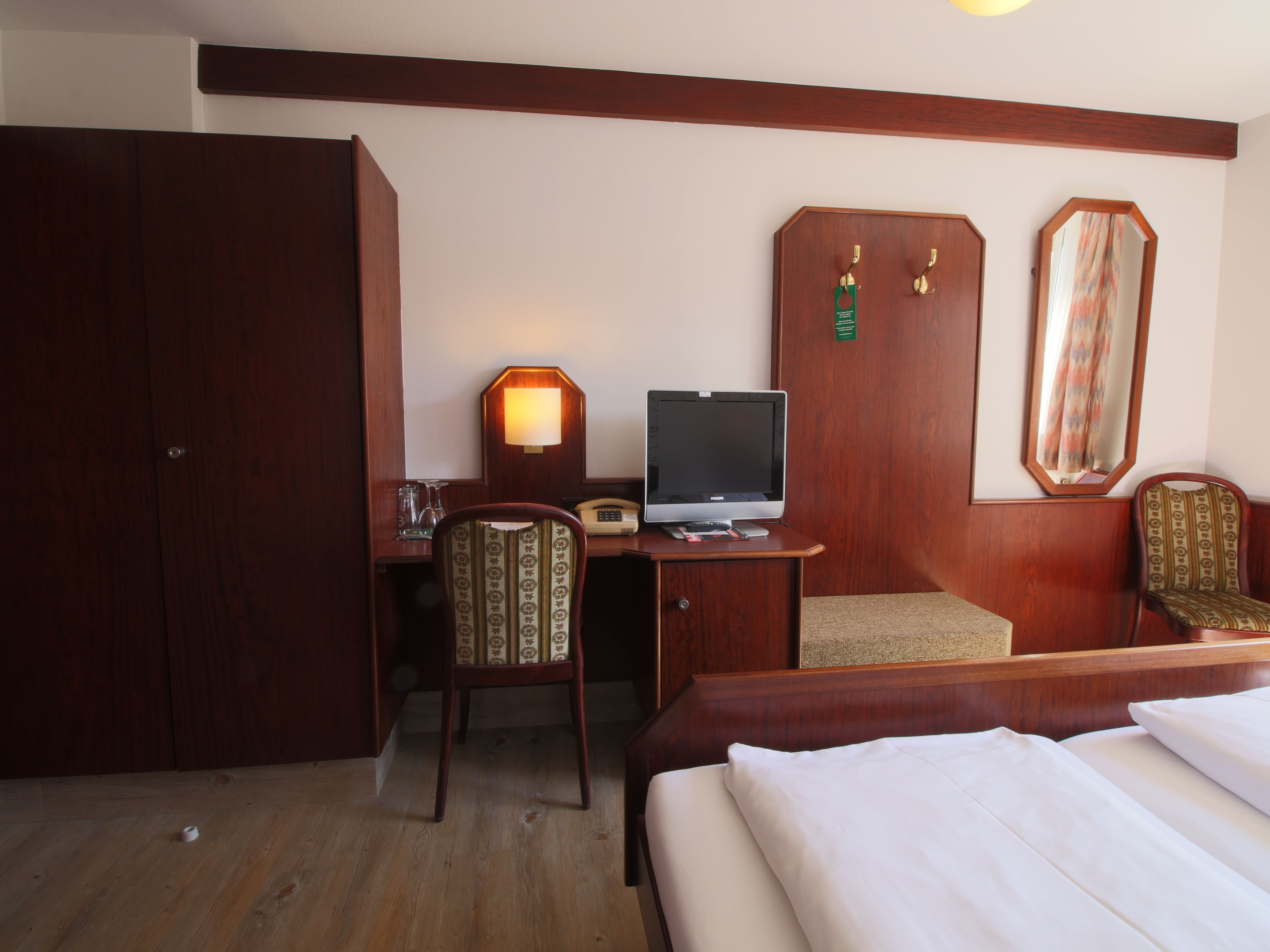 A hotel room with a bed, a desk with a computer, a chair, a wardrobe, and a mirror.