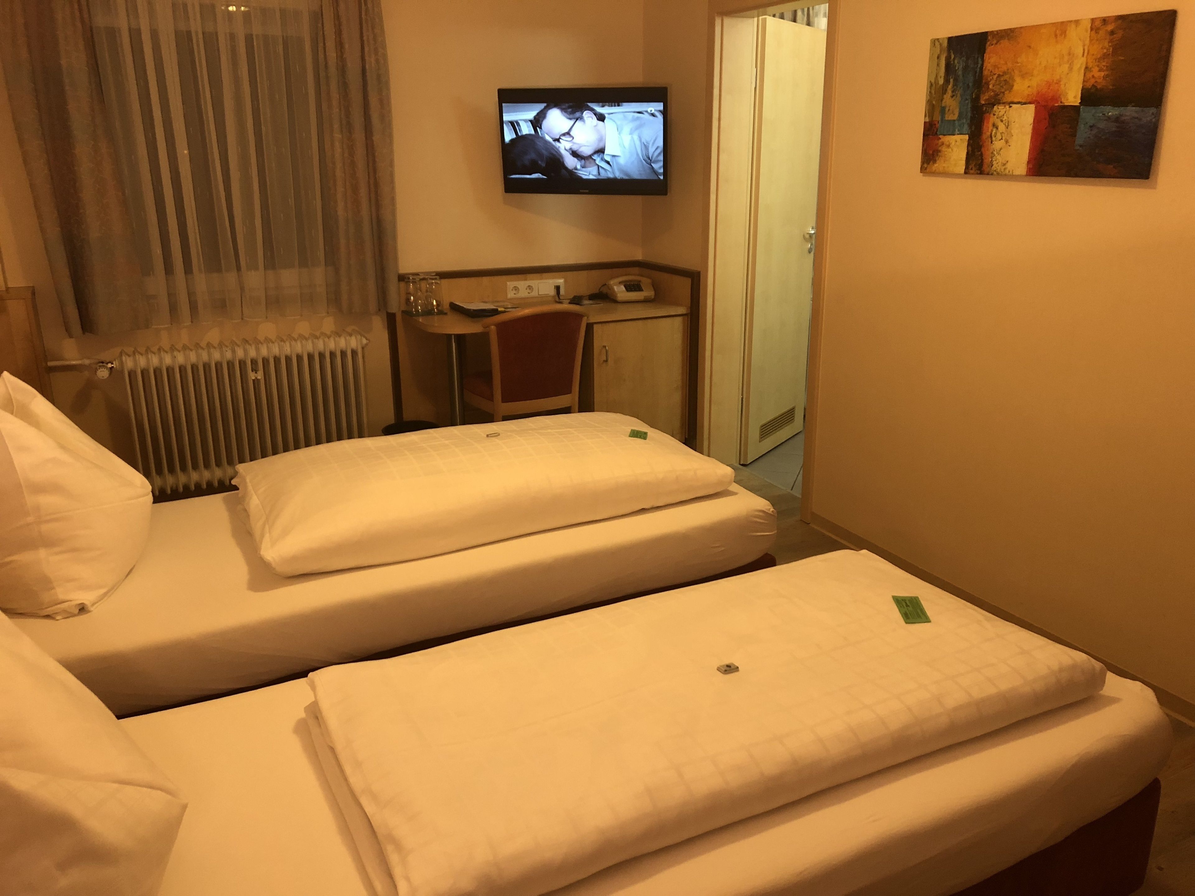 A hotel room with two beds, a desk, a chair, a radiator, a television, and a door.