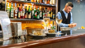 Una zona de bar con una variedad de botellas de licores en estantes, un barman preparando bebidas y una copa de vino sobre la barra.
