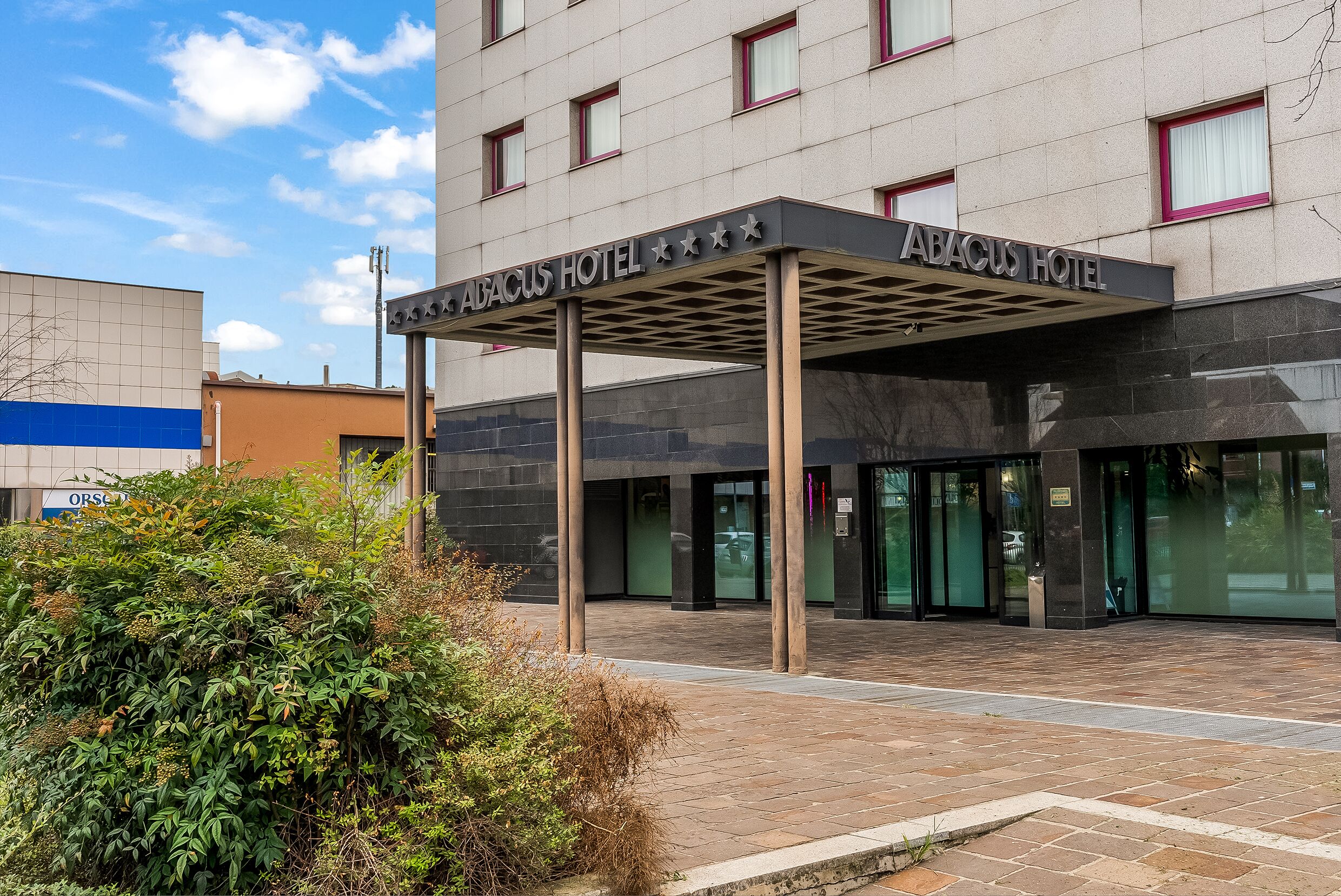 Exterior de l'Hotel Abacus, amb entrada coberta, façana moderna i zona enjardinada amb arbustos.