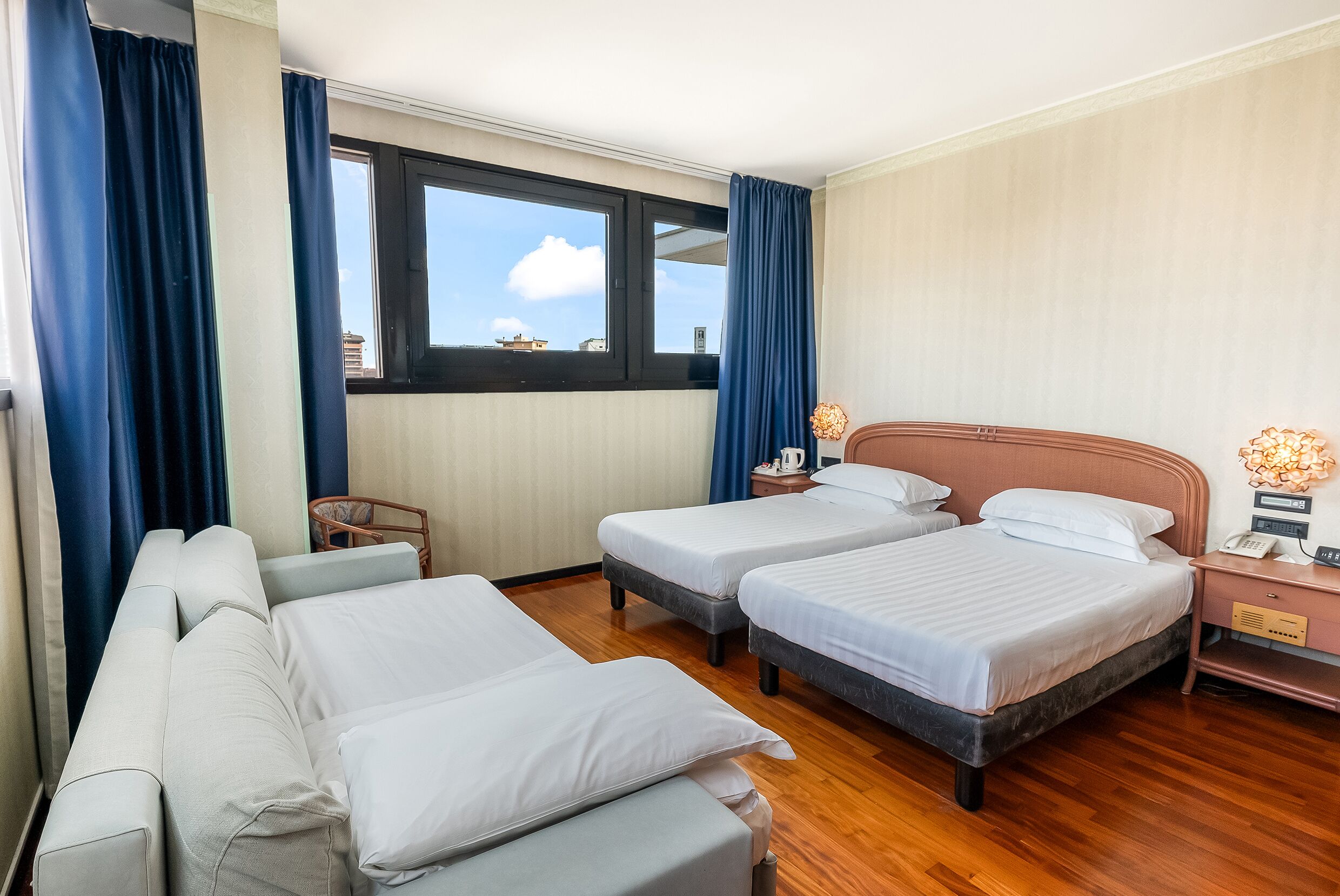 A hotel room with two single beds, a sofa, a wooden floor, a window with curtains, and a nightstand with a lamp.
