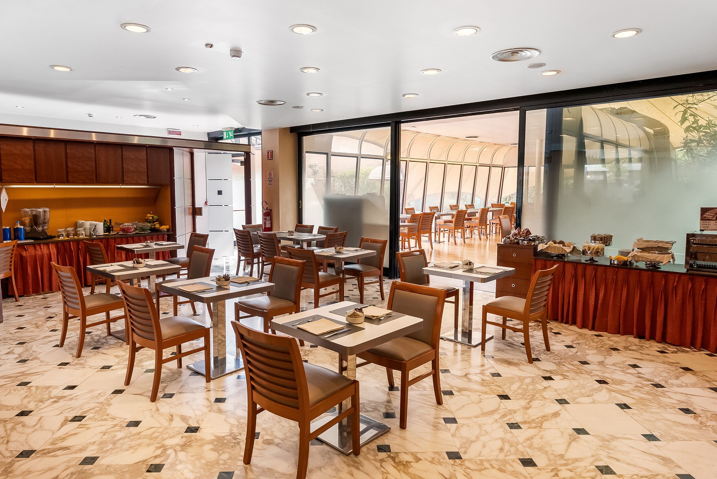 Un restaurante espacioso con suelo de mármol, sillas y mesas de madera. Cuenta con ventanas grandes y un separador de vidrio.