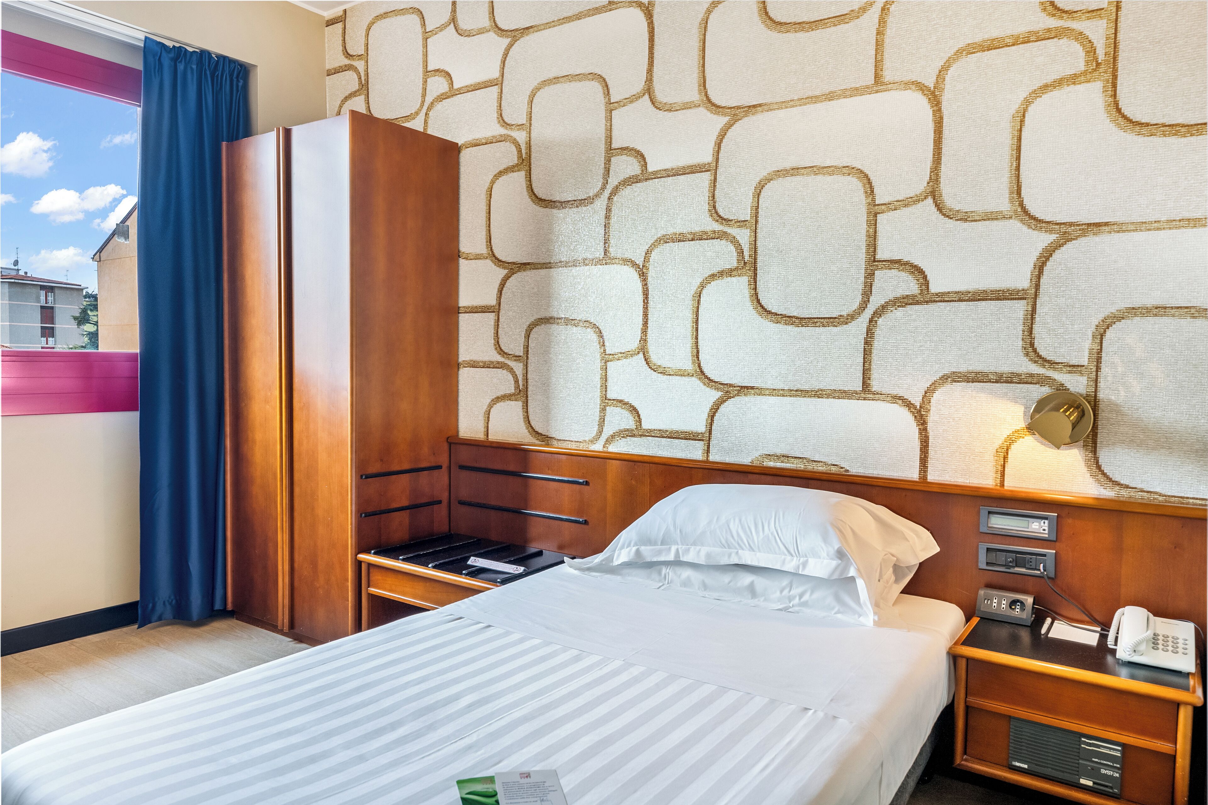 A hotel room with a bed, a wooden wardrobe, a nightstand with a phone, and a patterned wall.