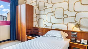 A hotel room with a bed, a wooden wardrobe, a nightstand with a phone, and a patterned wall.
