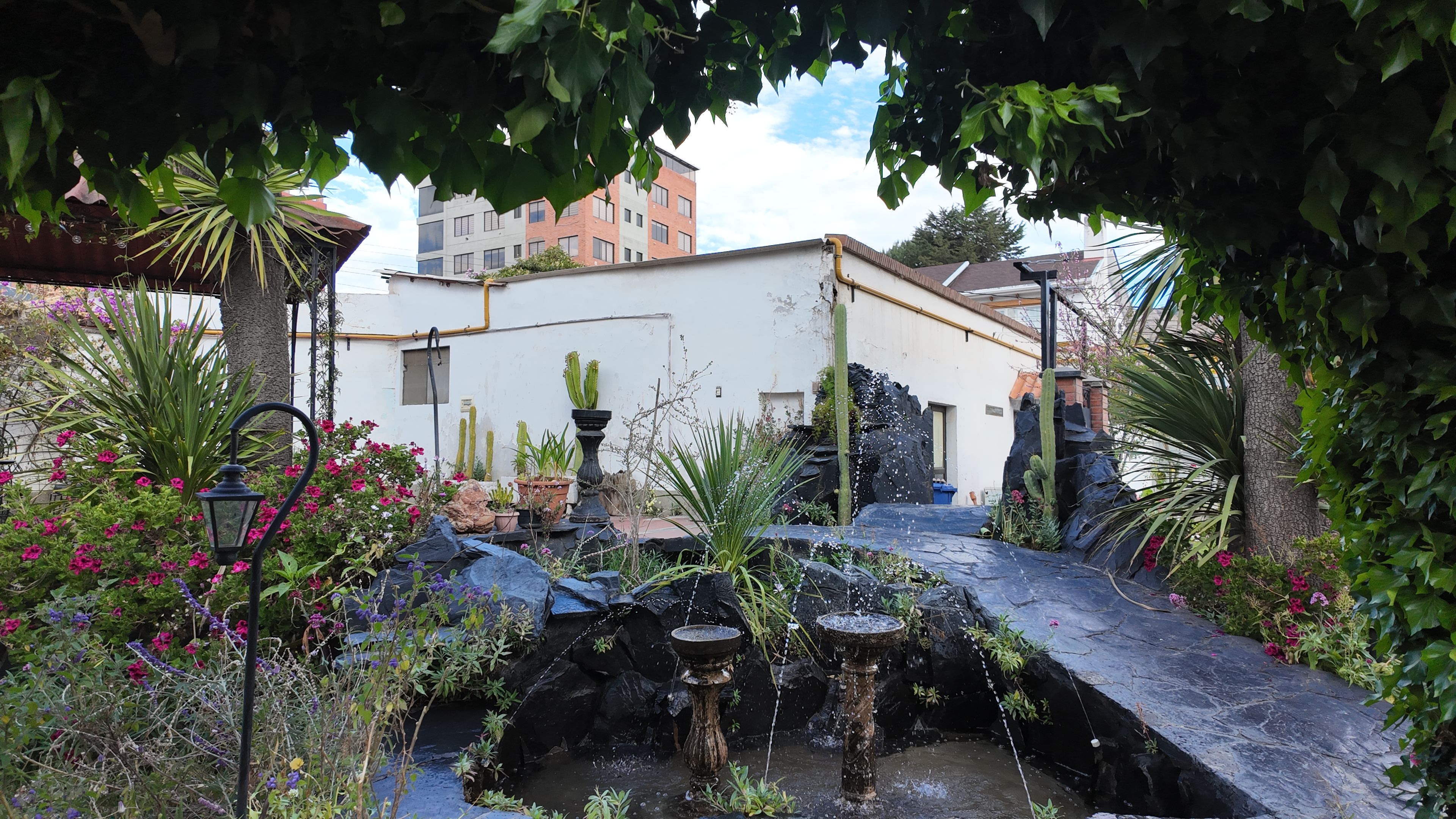 Un jardín con una fuente de agua, plantas en macetas y un edificio al fondo.