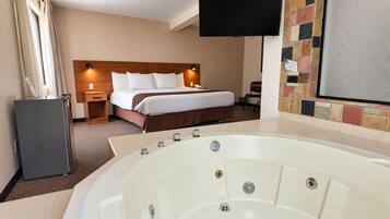 A hotel room with a large bathtub, a bed with white linens, a wooden headboard, a flat-screen TV mounted on the wall, and a small refrigerator.