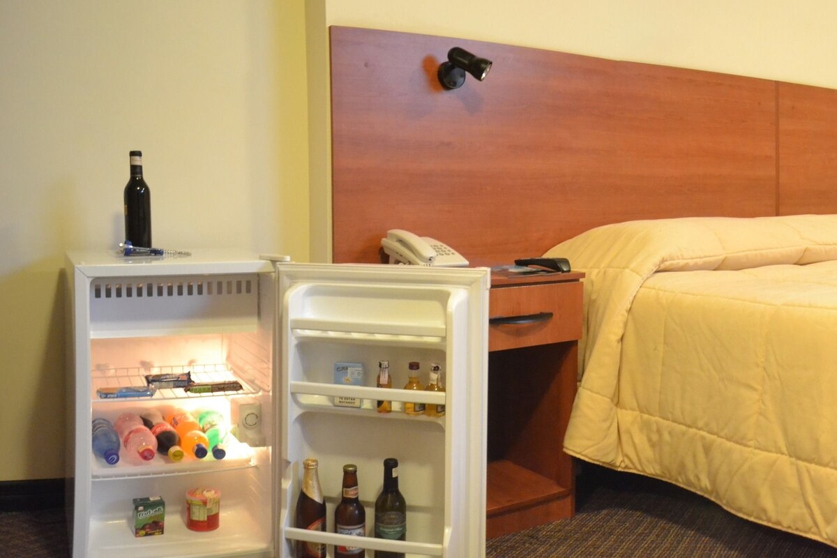 A hotel room with a bed, a desk, a chair, a refrigerator with beverages, and a phone.