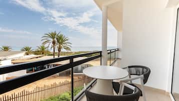 A balcony with a round table and two chairs, overlooking a beach with palm trees and a fence.