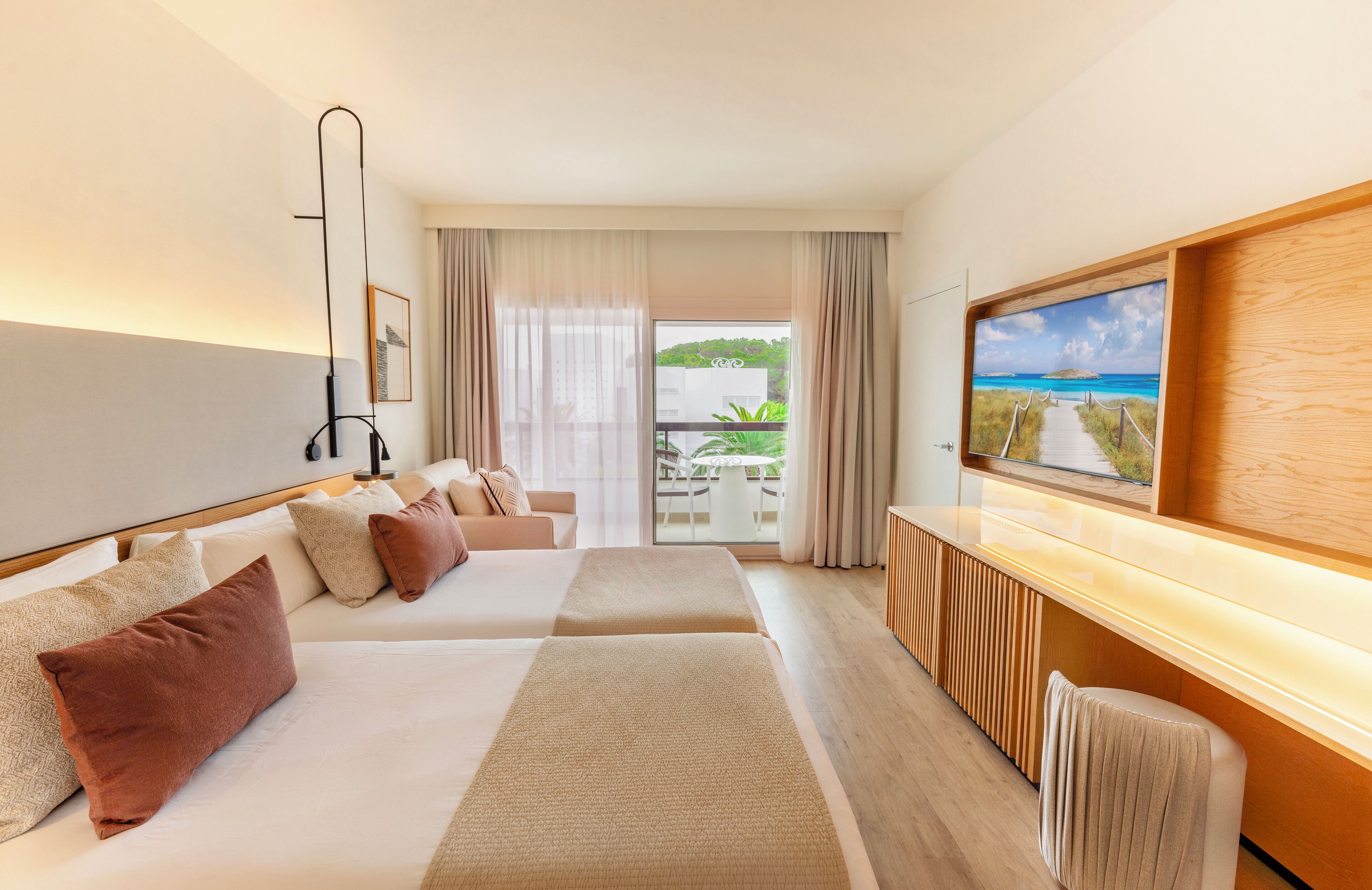 A hotel room with two beds, a sofa, a TV, and a balcony with a view of greenery and a beach.