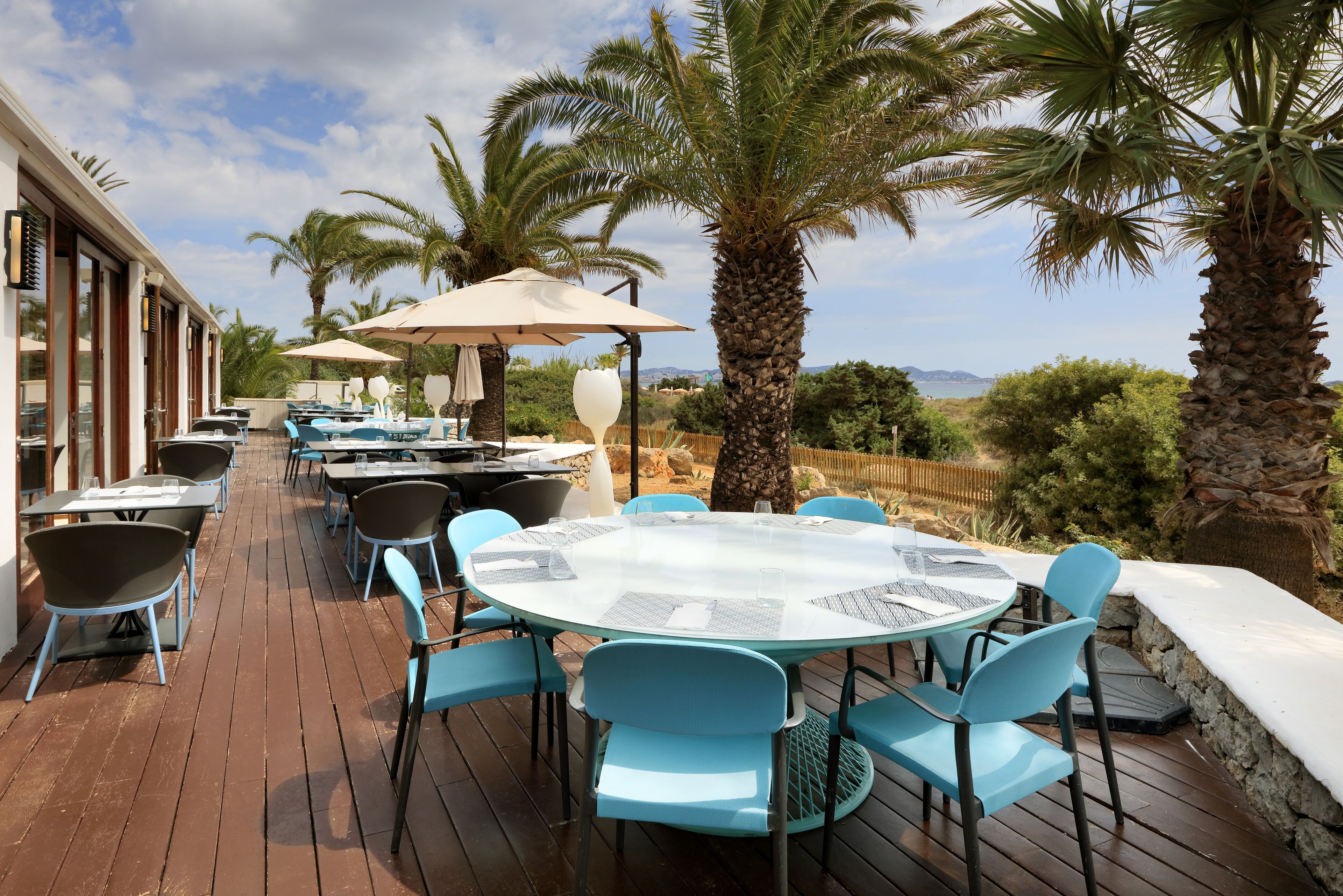 Un espacio de comedor al aire libre con mesas redondas y sillas azules, rodeado de palmeras y vistas al paisaje.