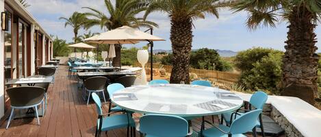 Un espacio de comedor al aire libre con mesas redondas y sillas azules, rodeado de palmeras y vistas al paisaje.