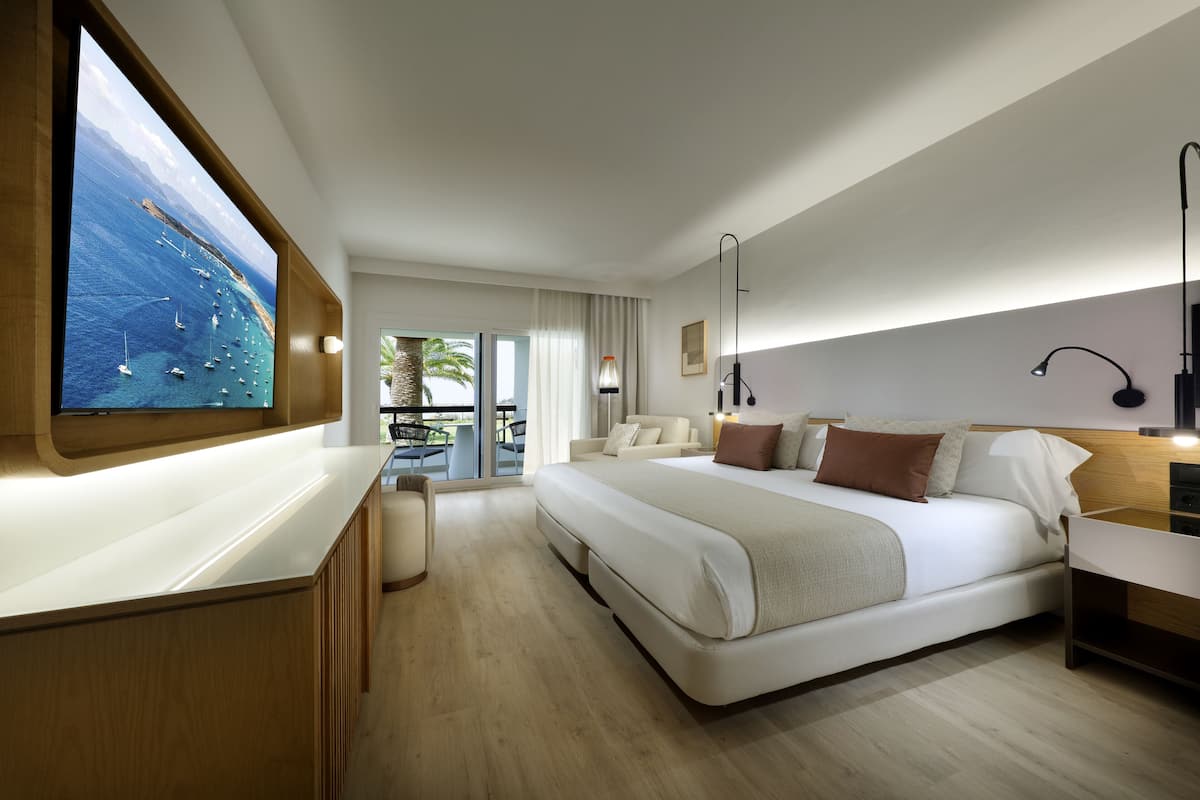 A modern hotel room with a large bed, a TV mounted on the wall, a balcony with a view of boats, and a wooden desk.