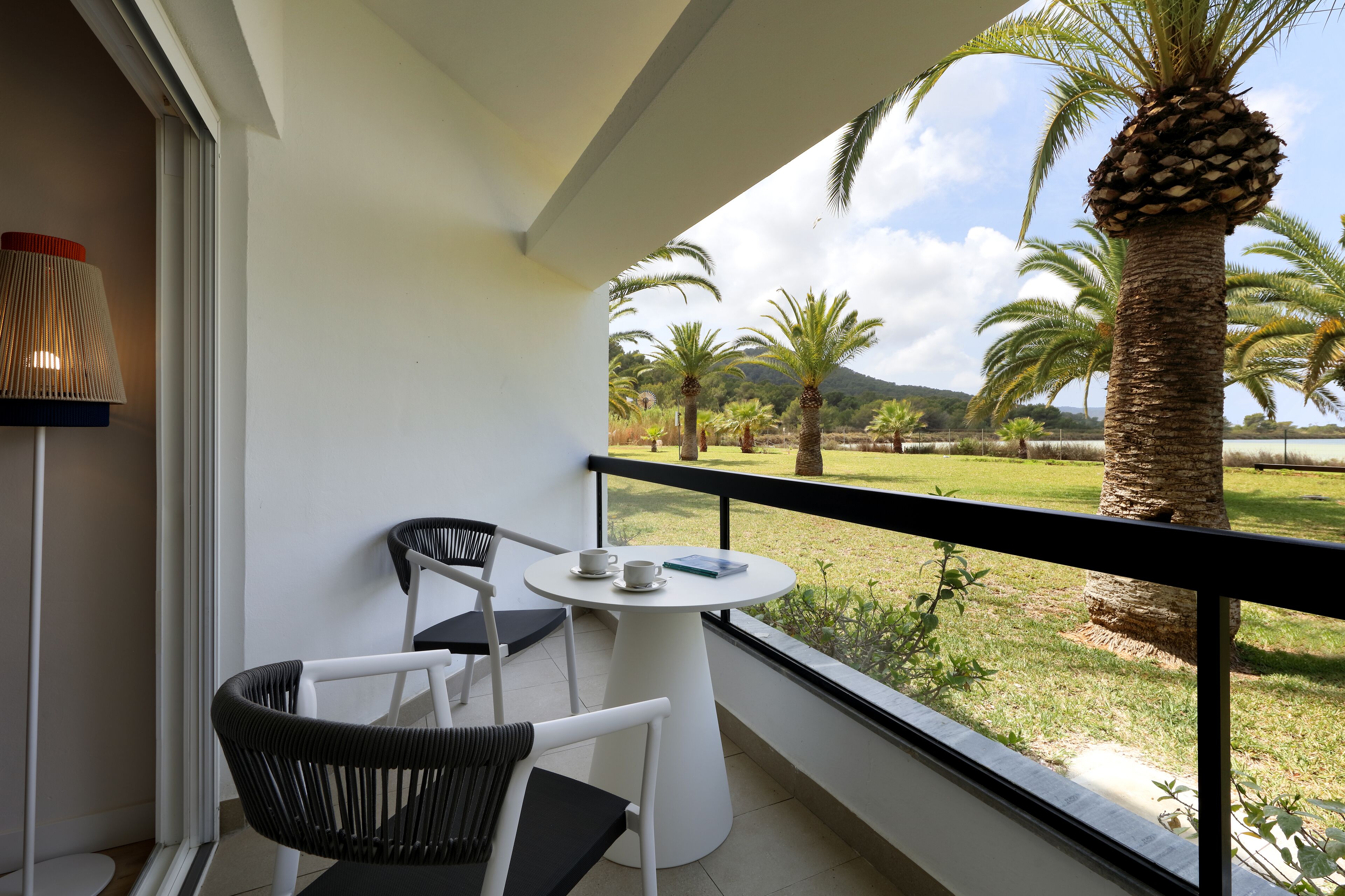A balcony with a white table and two chairs, overlooking a grassy area with palm trees and a mountain in the distance.