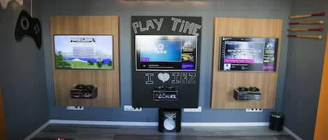 Een speelkamer met drie televisies die verschillende games tonen, een neonbord met de tekst 'The Black & White CLUB' en diverse spelaccessoires.