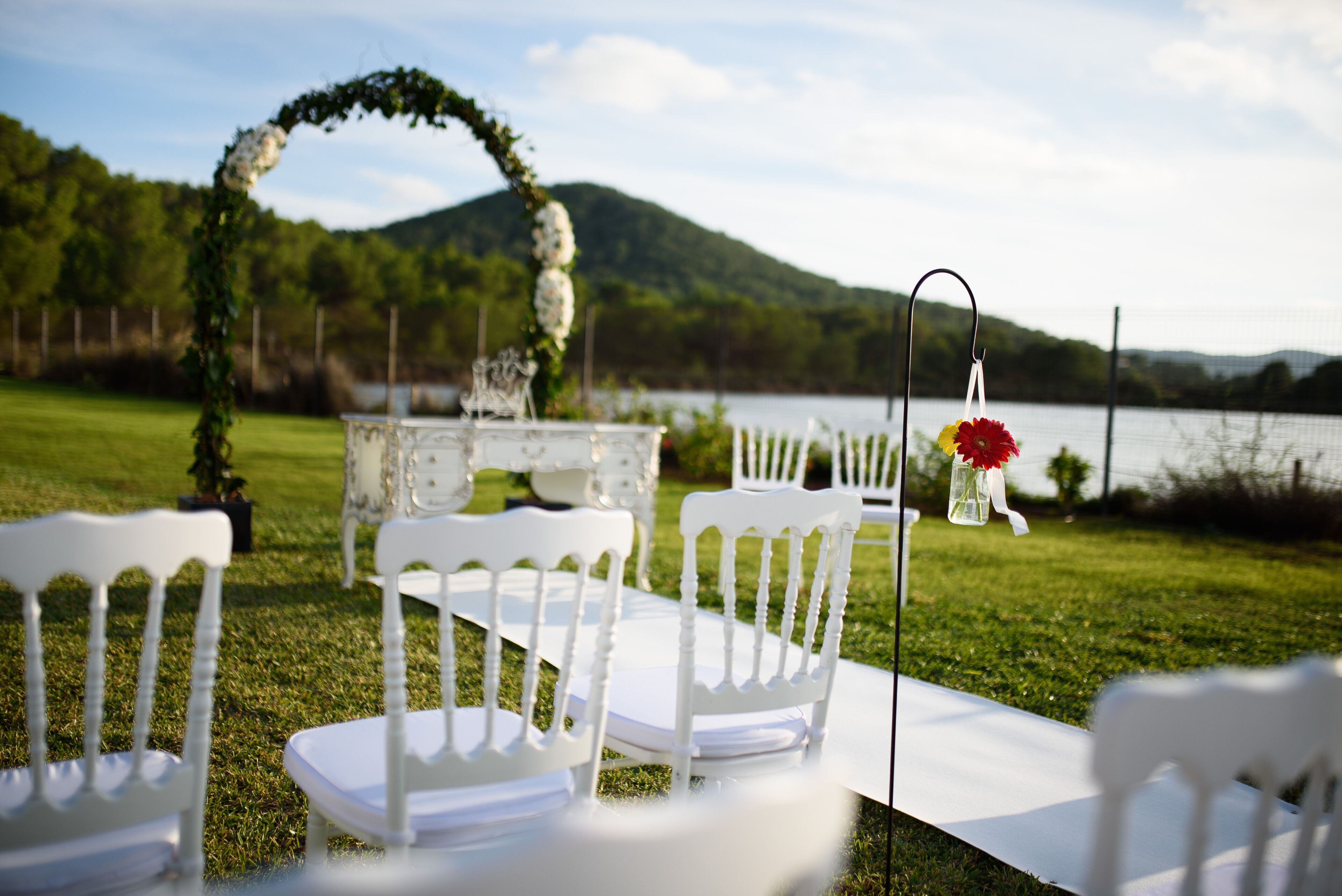 Outdoor wedding ceremony chairs arranged on a white tablecloth, a floral arch, and a lake view.