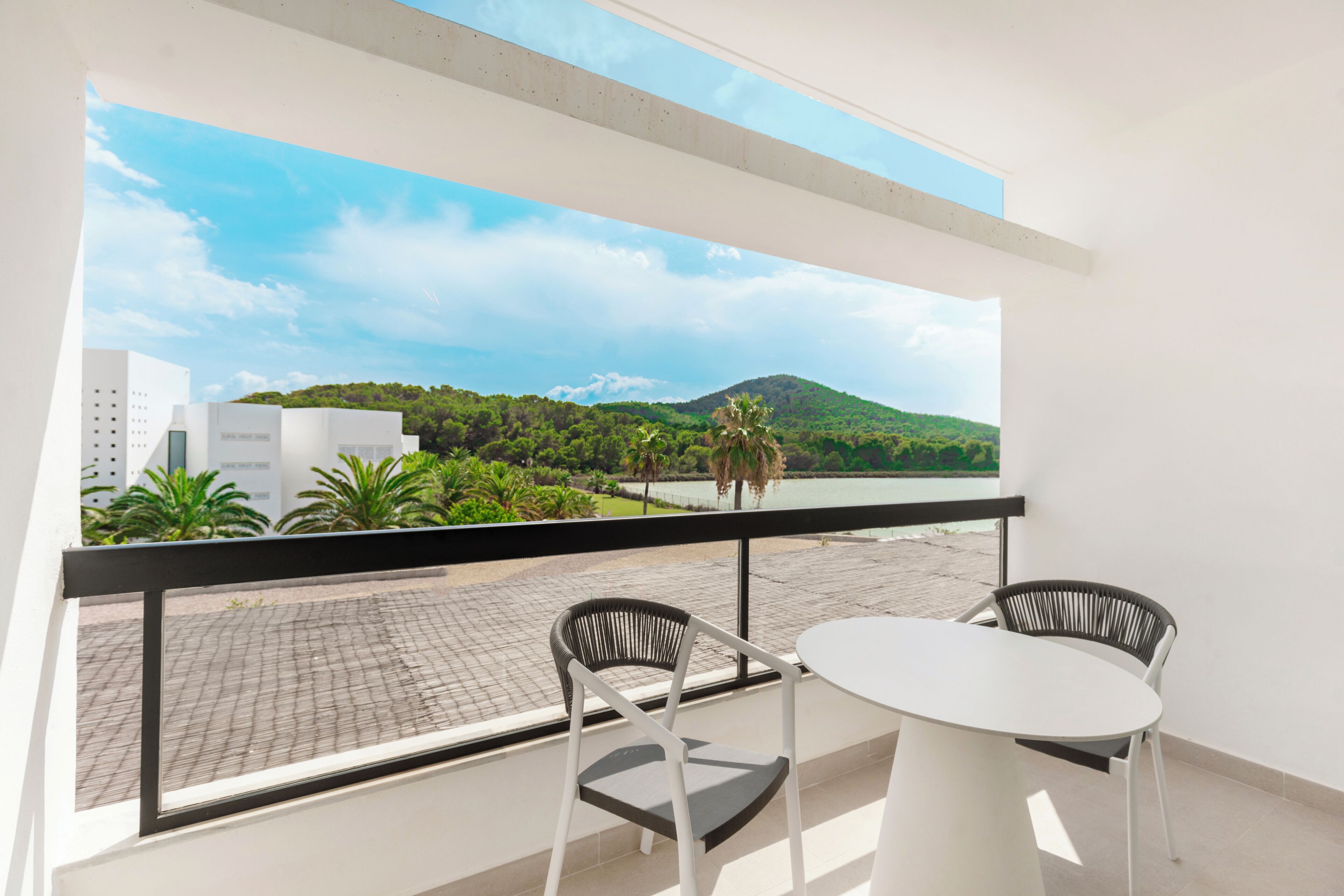 A balcony with a white table and two chairs, overlooking a paved area with greenery and a body of water, with buildings in the background.