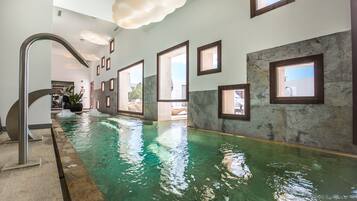 An indoor swimming pool with clear water, a modern design with stone walls and framed windows, and a ceiling with a cloud-like light fixture.