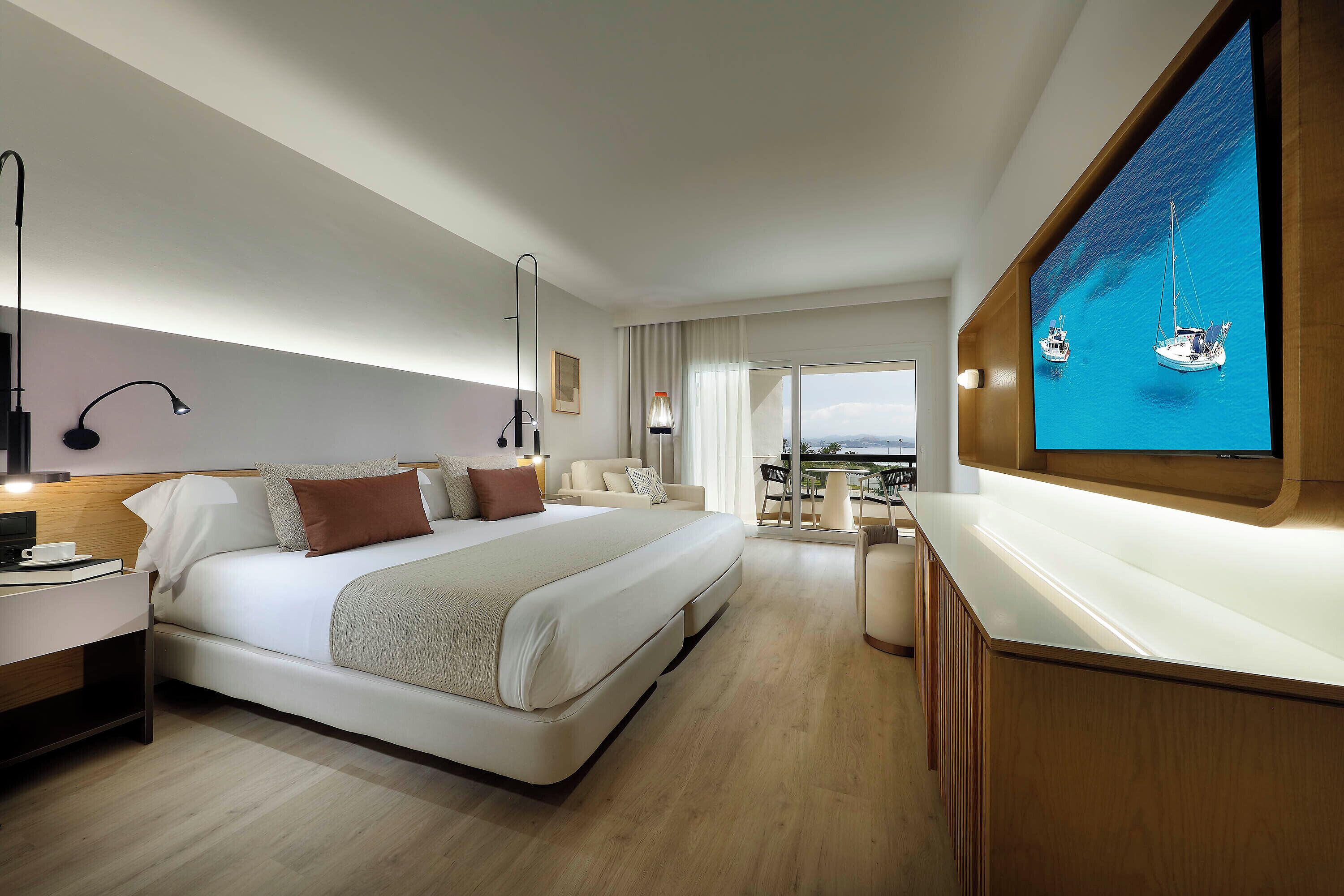 A modern hotel room with a large bed, bedside tables, a TV, and a view of the sea.