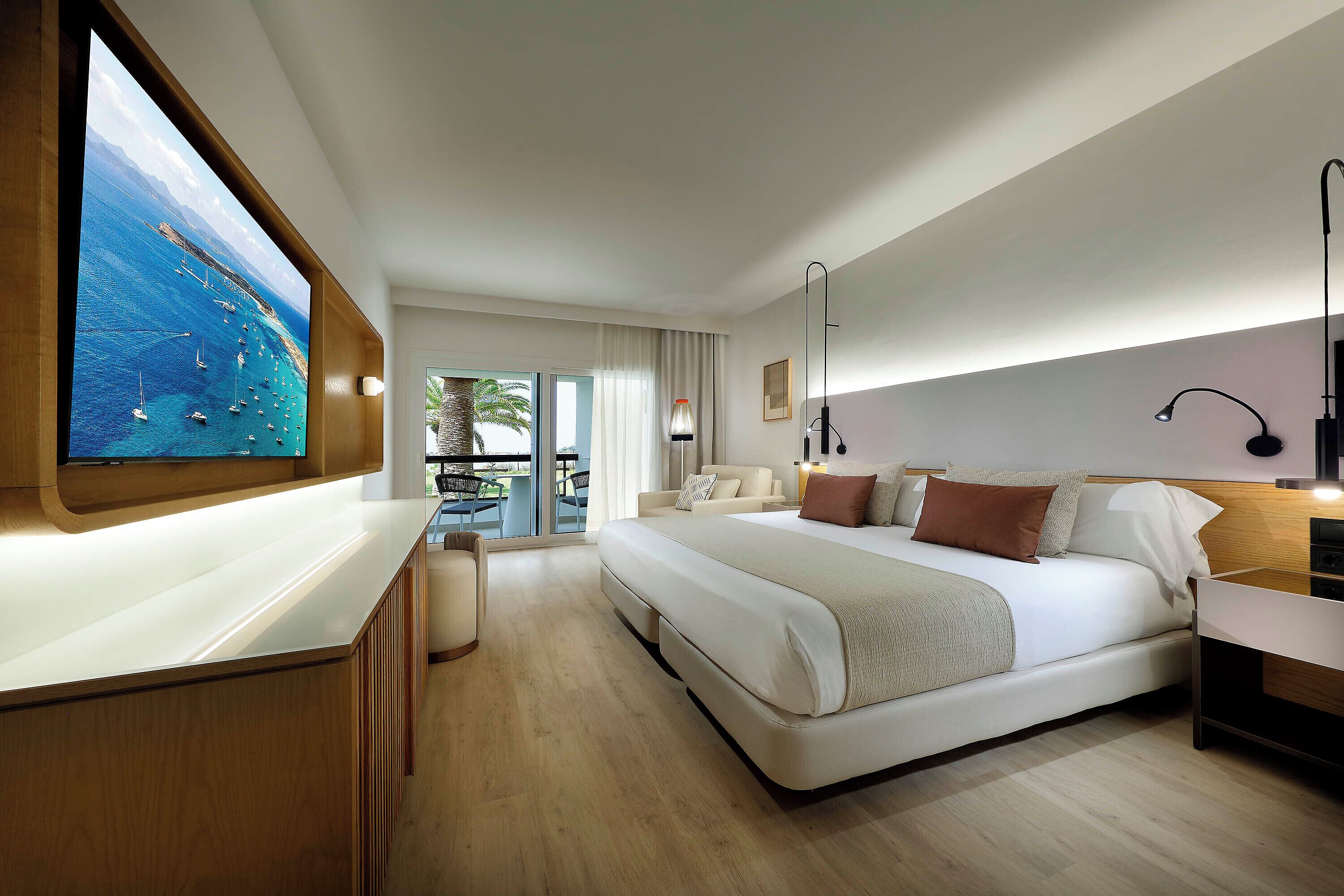 A modern hotel room with a large bed, a TV mounted on the wall, a balcony with a view of the sea, and a wooden desk.