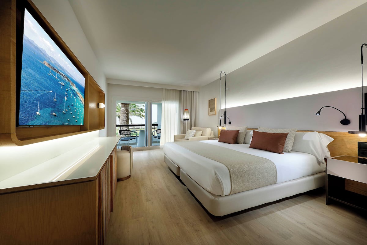 A modern hotel room with a large bed, a TV mounted on the wall, a balcony with a view of the sea, and a wooden desk.