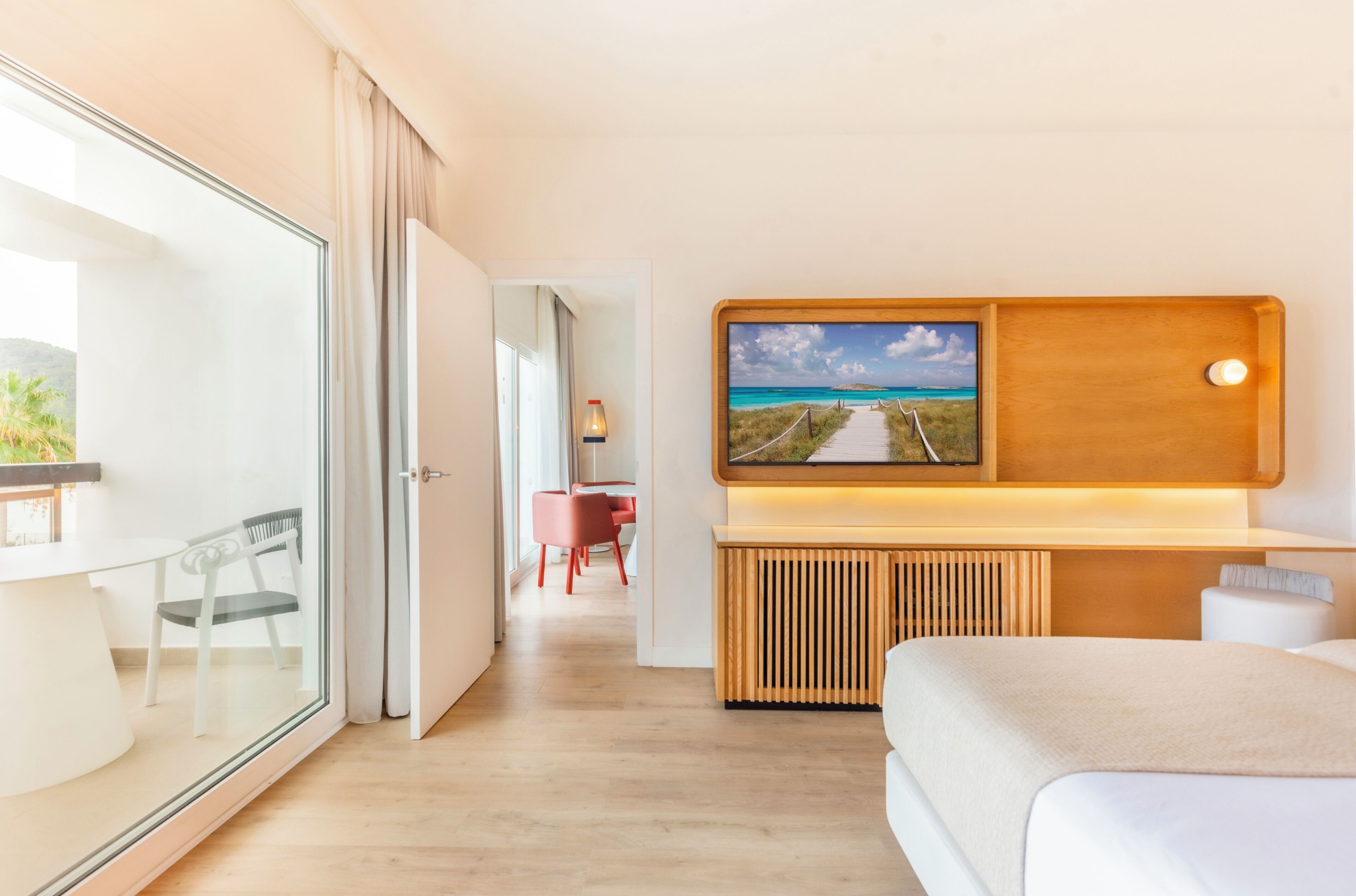A modern hotel room with a glass door leading to a balcony, a wooden bedside table, a bed, and a wall-mounted TV displaying a scenic view.