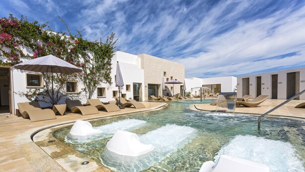 Un área de piscina con agua cristalina, sillas de descanso y un edificio moderno con grandes ventanales.