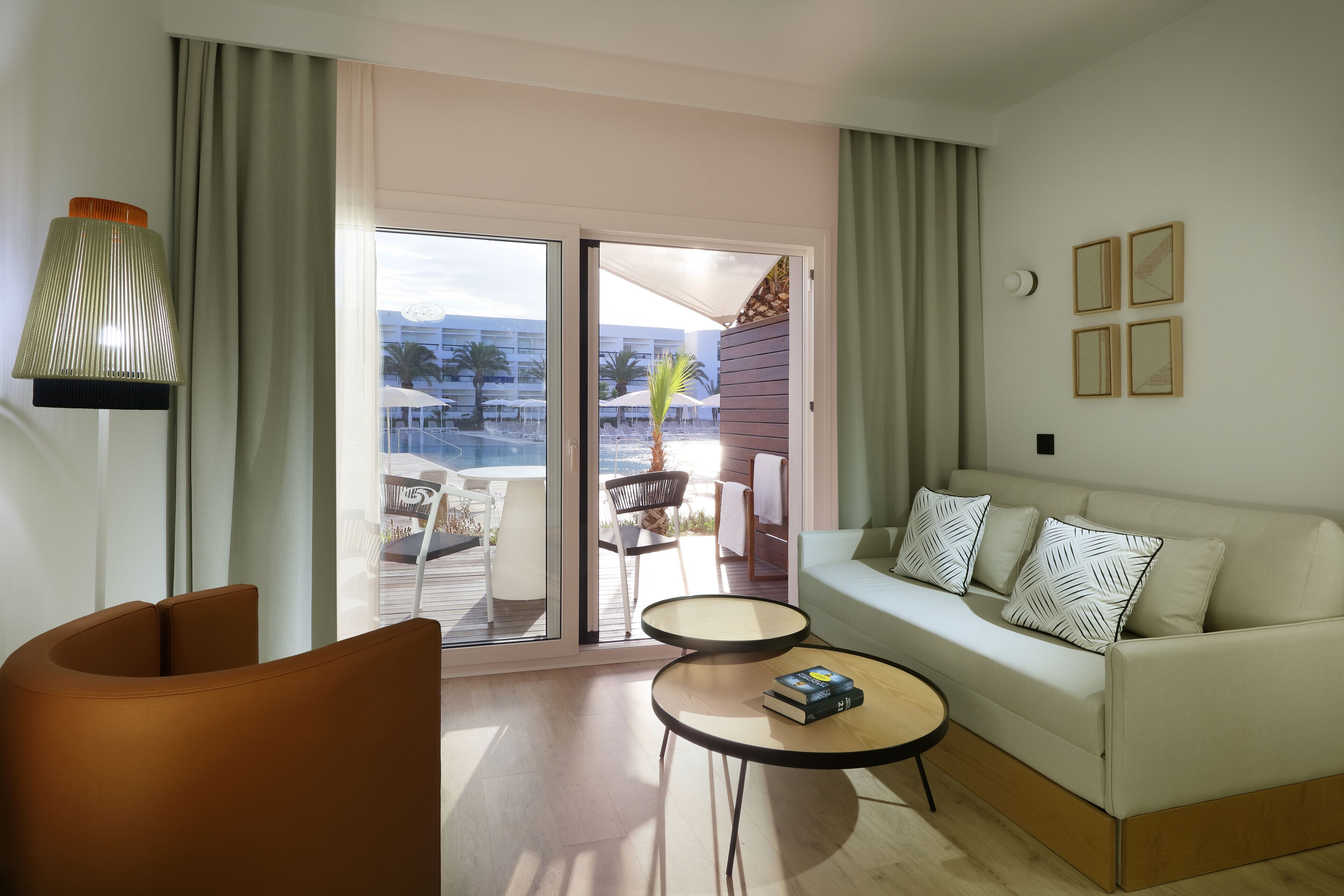 A modern living room with a sofa, two side tables, a lamp, and a view of an outdoor deck with chairs and umbrellas.