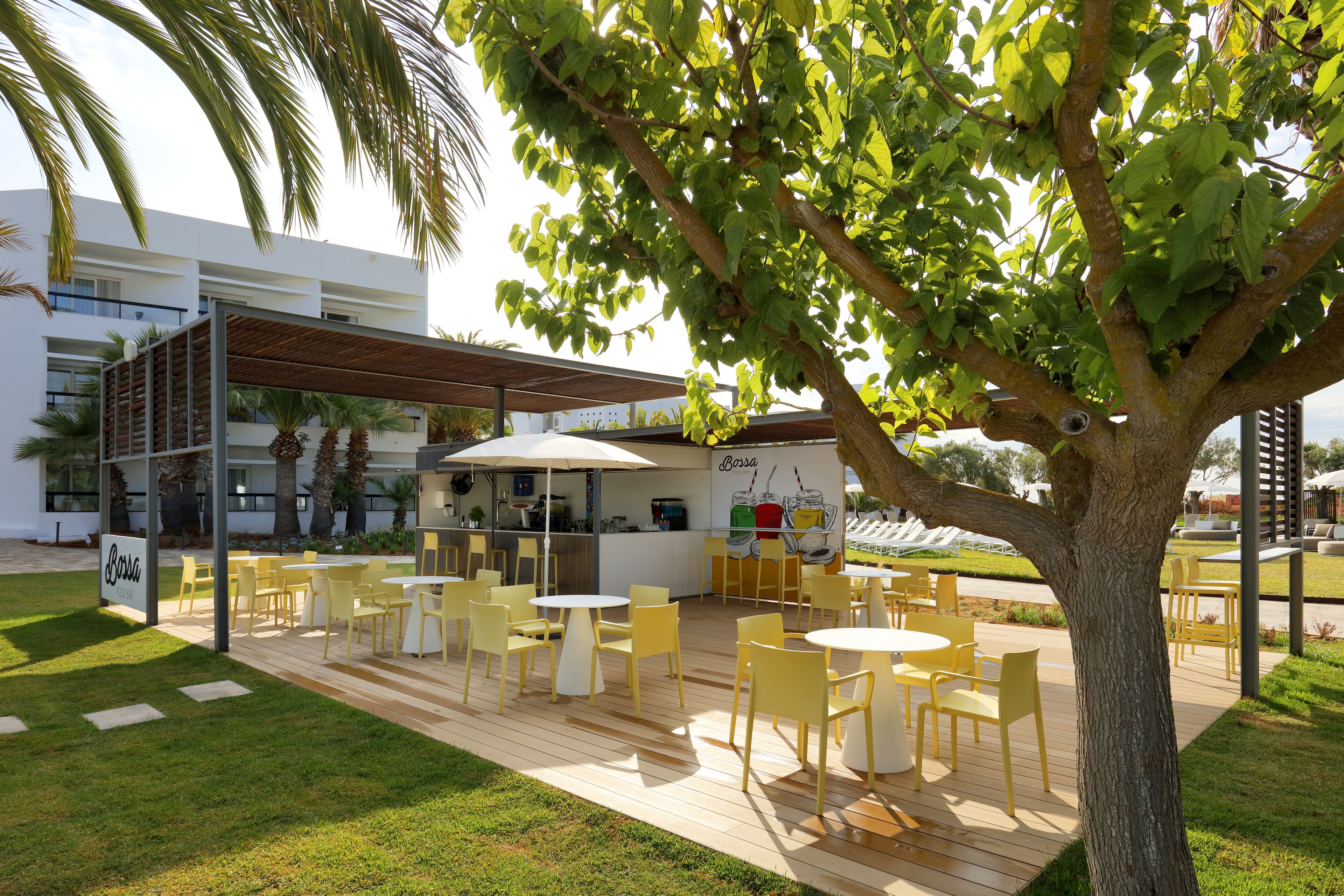An outdoor seating area with yellow chairs and tables, a covered section, and a tree in the foreground.