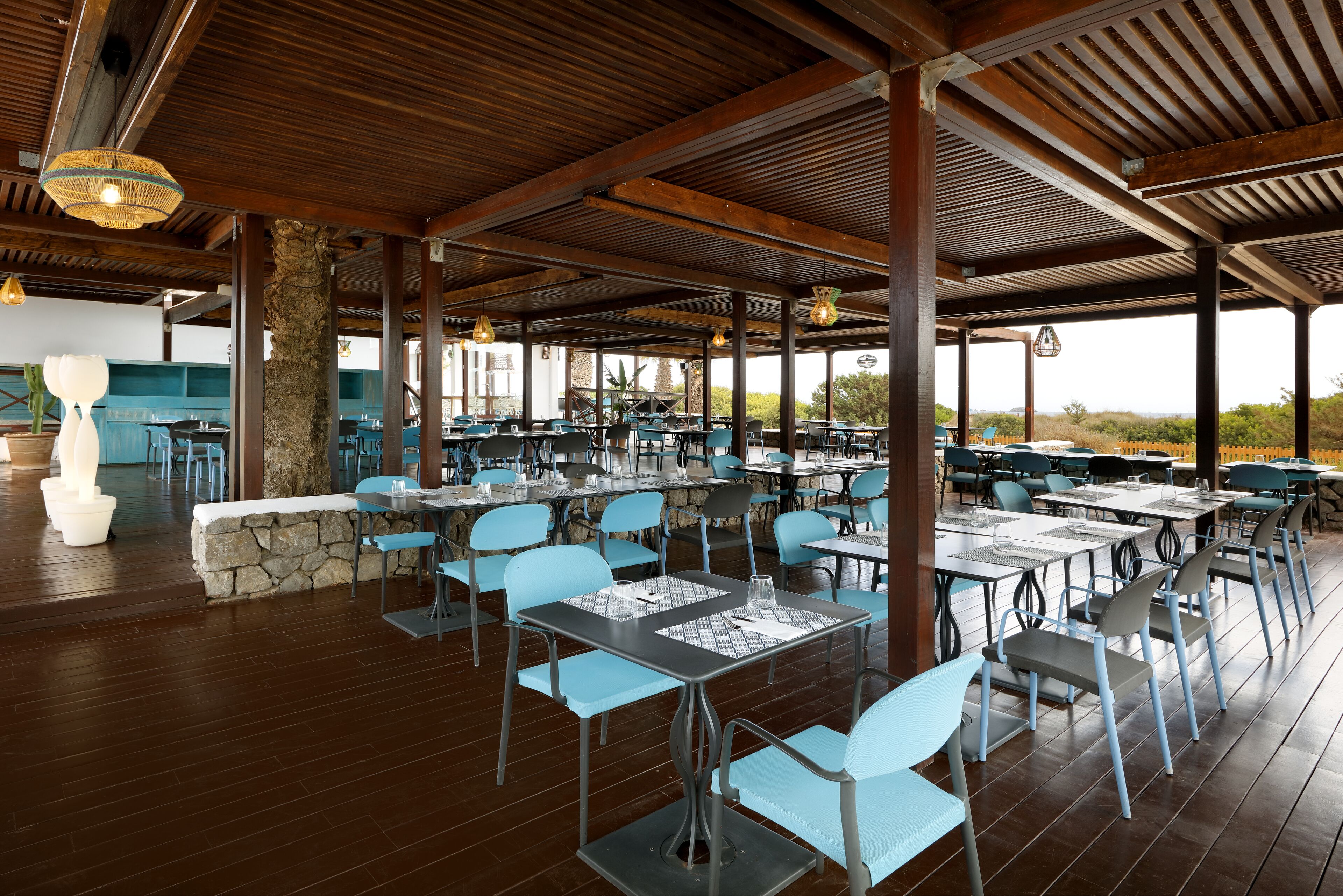 An outdoor dining area with wooden beams, stone pillars, and blue chairs. Tables are set with placemats and glasses.