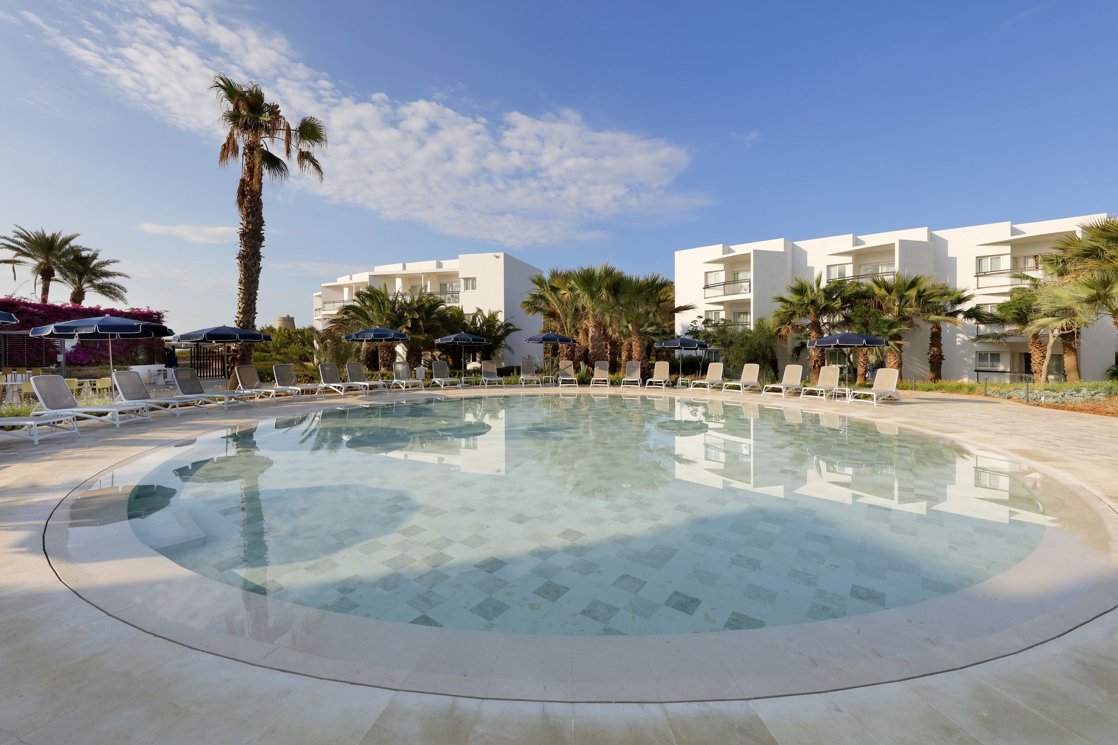 A pool area with lounge chairs and umbrellas, surrounded by palm trees and modern buildings.