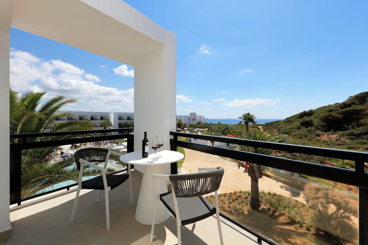 A balcony with a white table and two chairs, overlooking a beach and buildings.