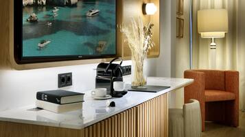 A modern hotel room with a large flat-screen TV displaying a coastal scene, a coffee machine, a vase with dried plants, and a comfortable orange chair.