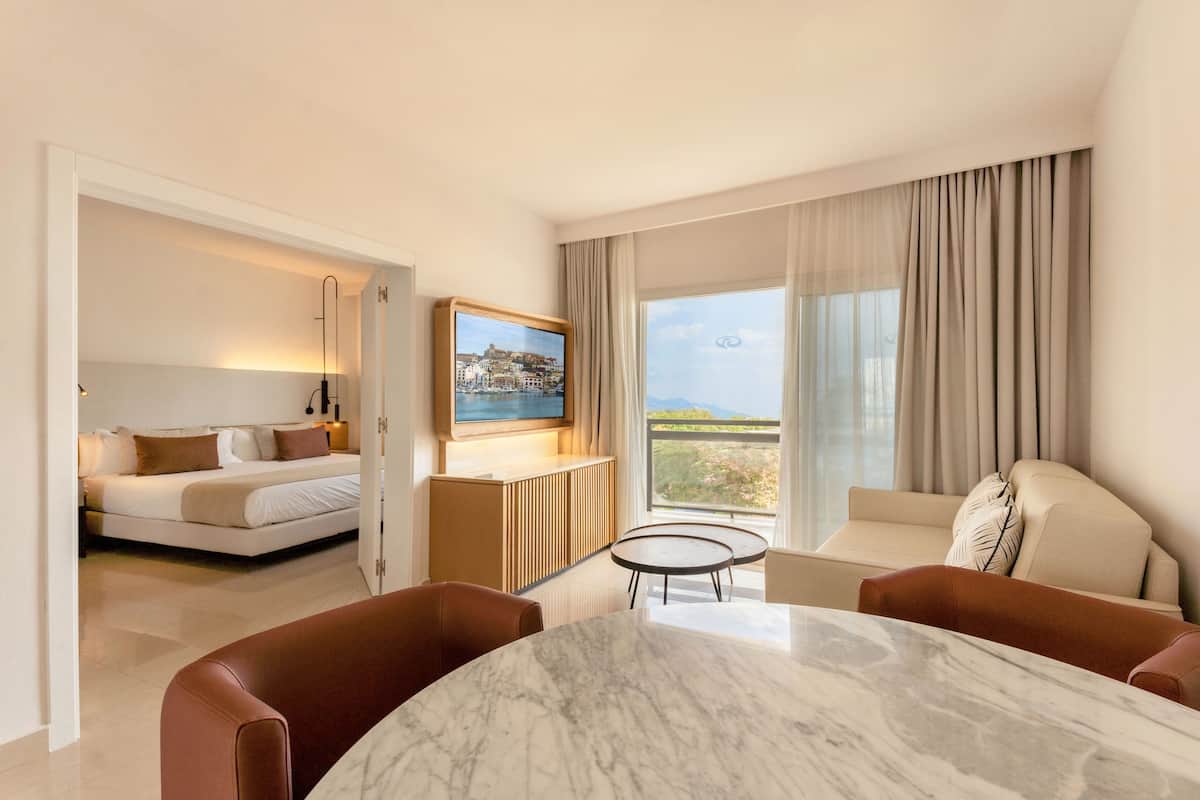 A hotel room with a bed, a seating area with a table, a television mounted on the wall, and a large window with curtains.