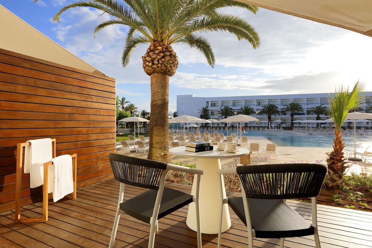 A wooden deck with two chairs and a table, overlooking a pool area with umbrellas and lounge chairs.
