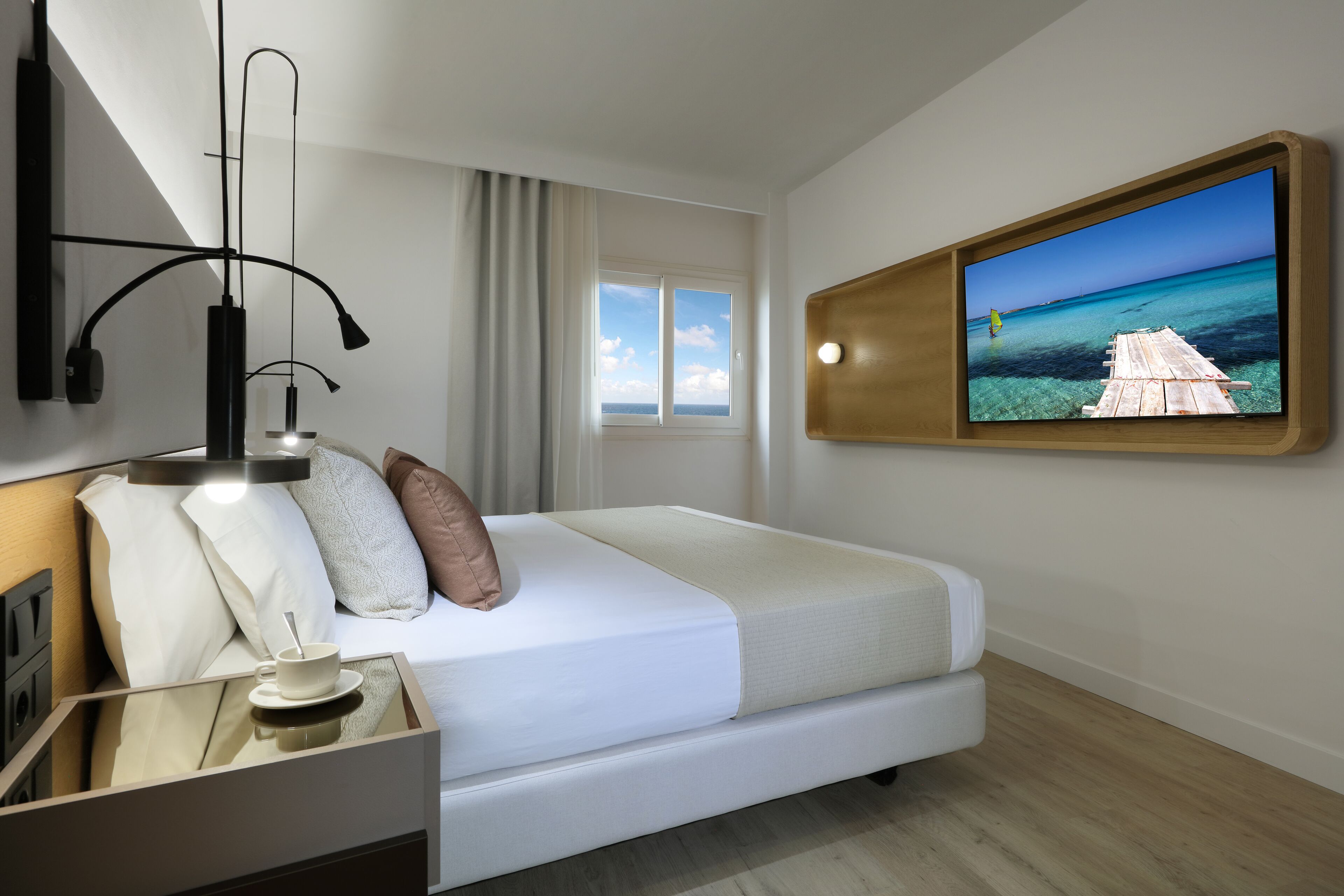 A modern hotel room with a bed, a bedside table with a cup and saucer, a wall-mounted TV displaying a beach scene, and a window with curtains.