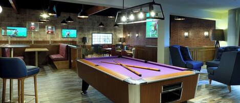 A pool table with cue sticks, a bar area with stools, and multiple televisions mounted on the wall.