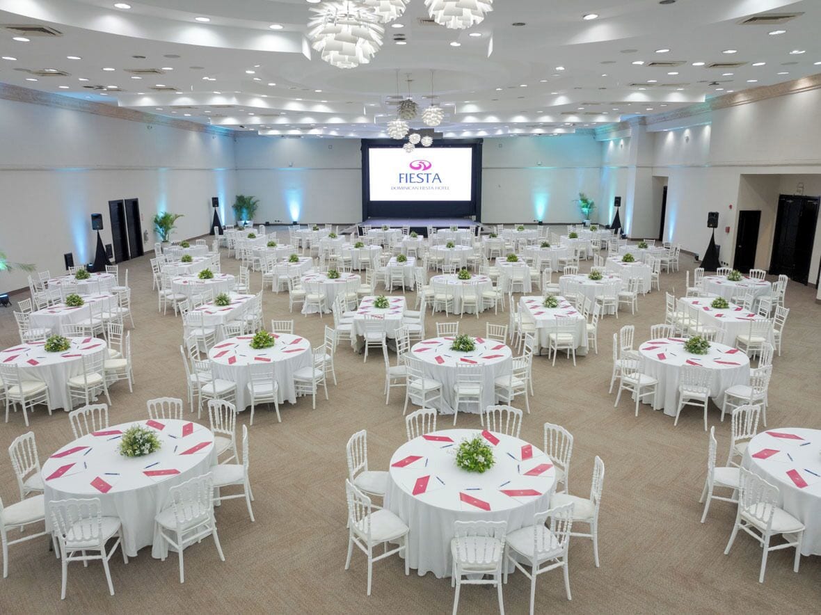 A large conference room with multiple round tables, each with white chairs and centerpieces. A large screen at the front displays the text "FIESTA."