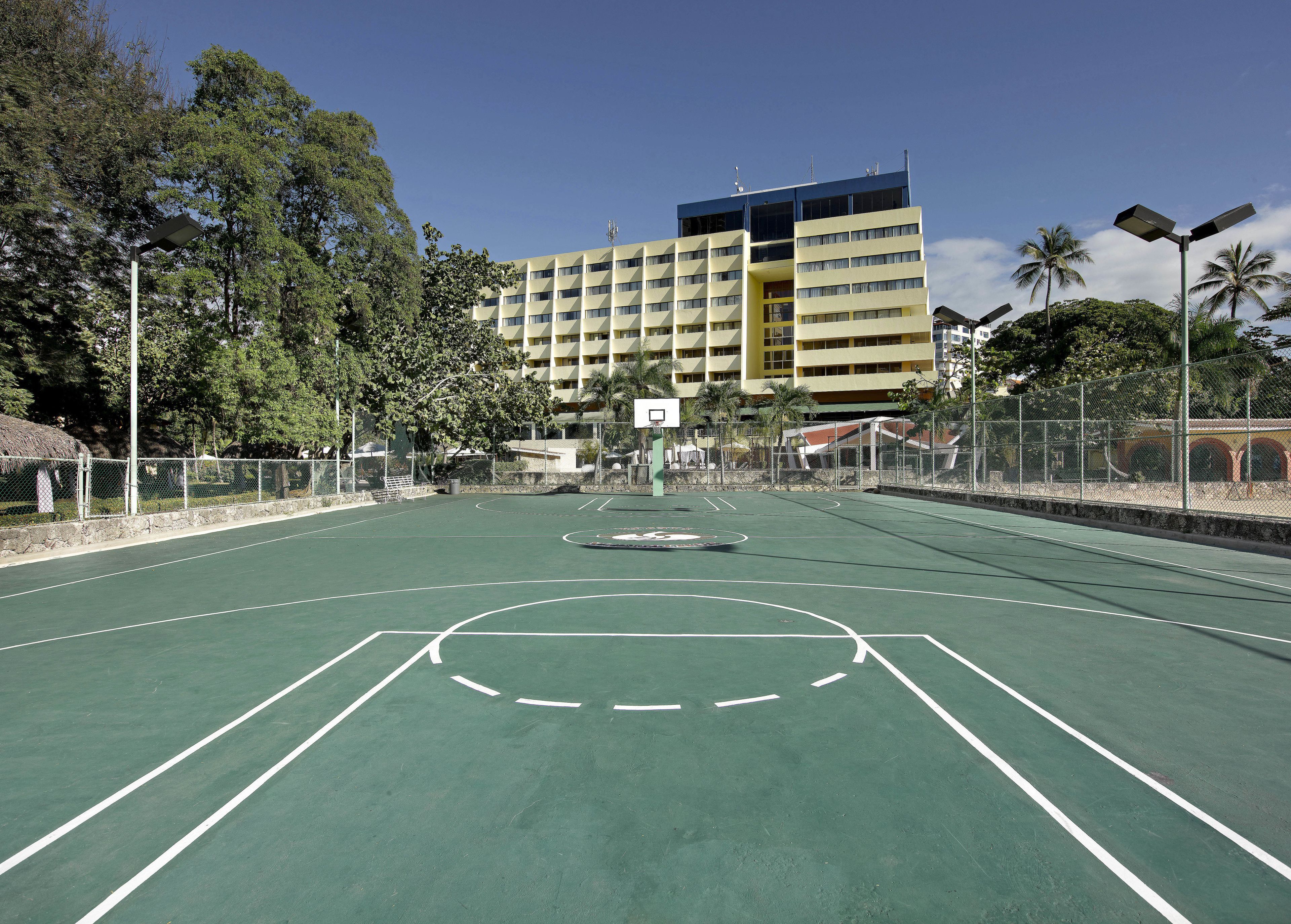Una cancha de baloncesto al aire libre con aro, rodeada por una valla, y un edificio de varios pisos al fondo.