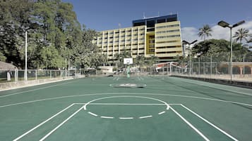 An outdoor basketball court with a net, surrounded by a fence, and a multi-story building in the background.