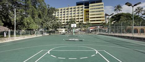 An outdoor basketball court with a net, surrounded by a fence, and a multi-story building in the background.