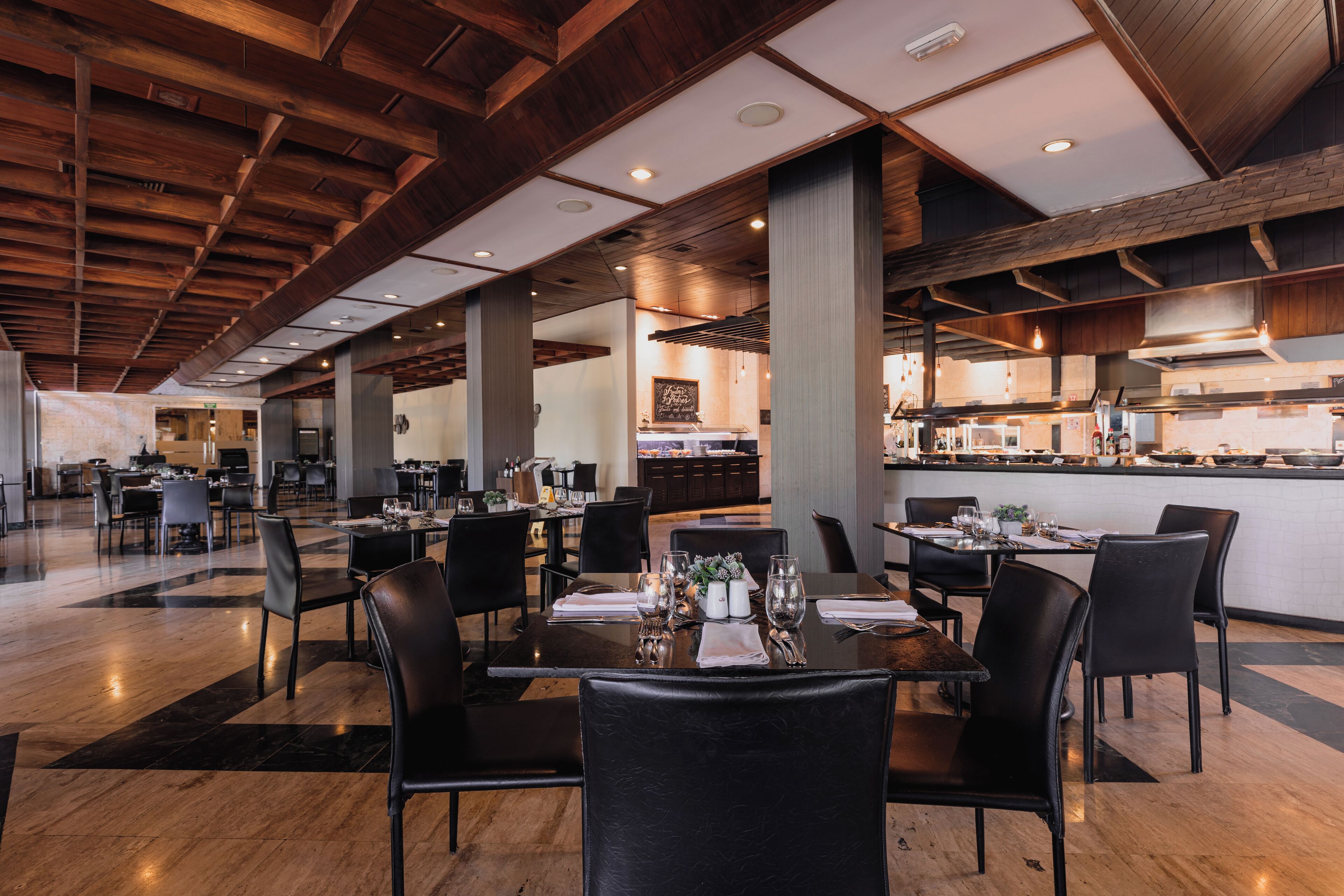 A spacious restaurant with high ceilings, wooden beams, and a central kitchen area. Tables are set with white tablecloths and black chairs.